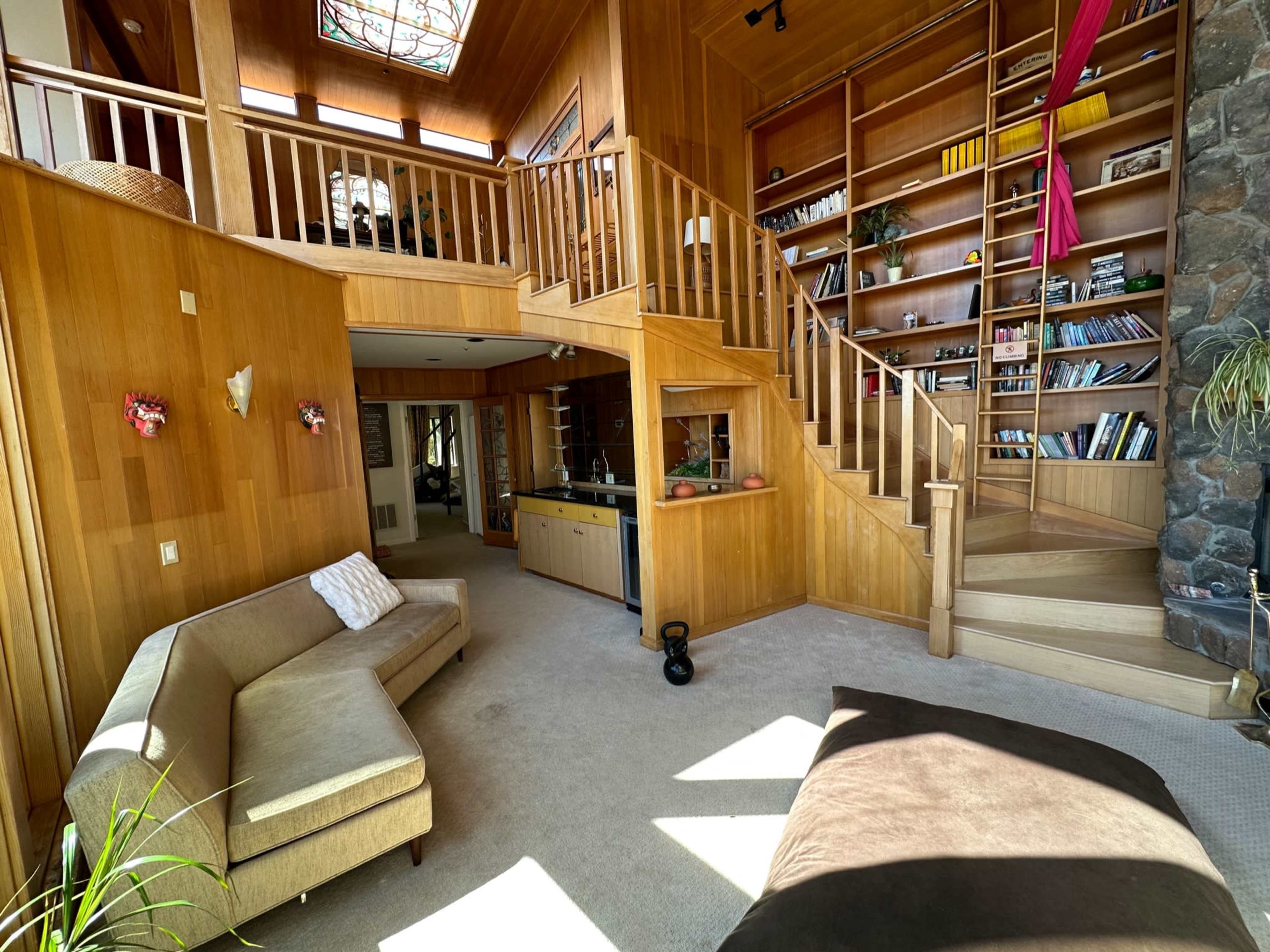 The image shows a wooden interior space with a staircase leading to an upper level, a cozy seating area, and a stone wall adorned with shelves filled with books and decorative items.