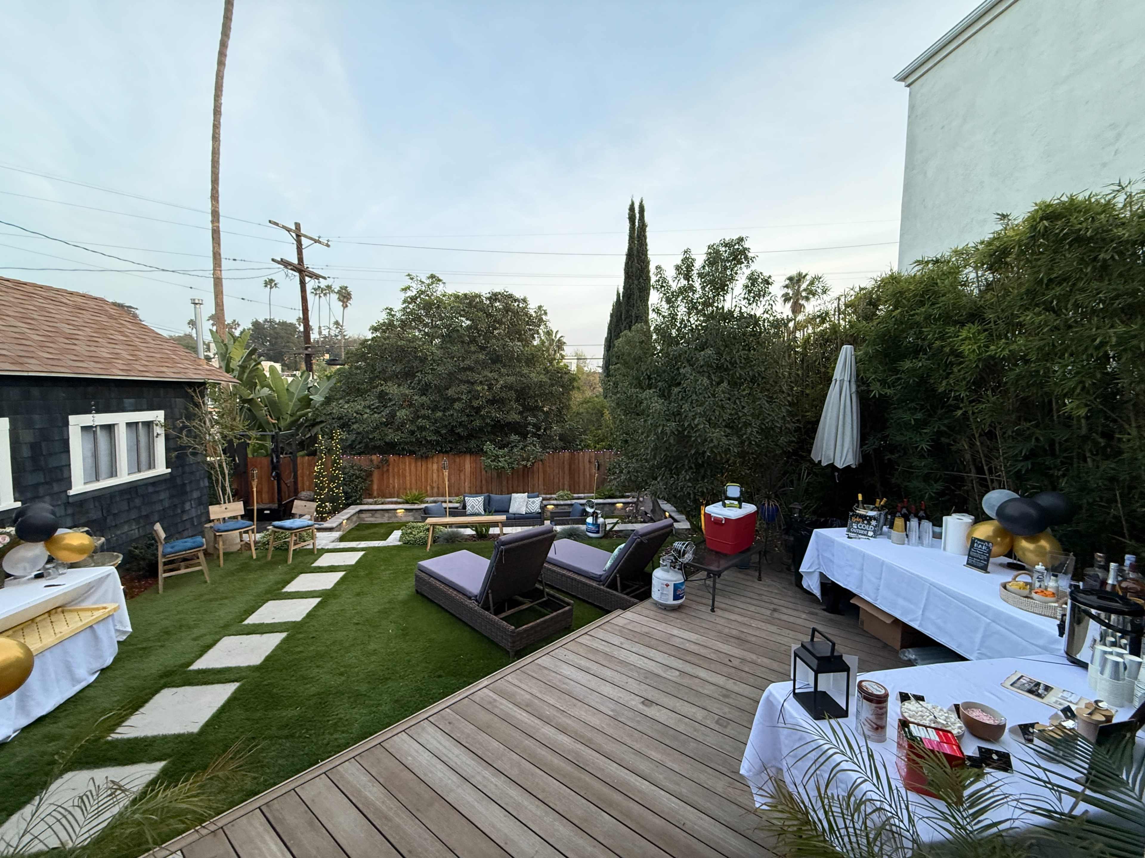 The image shows a backyard event space featuring lounge chairs, a bar setup, and decorative tables surrounded by greenery.
