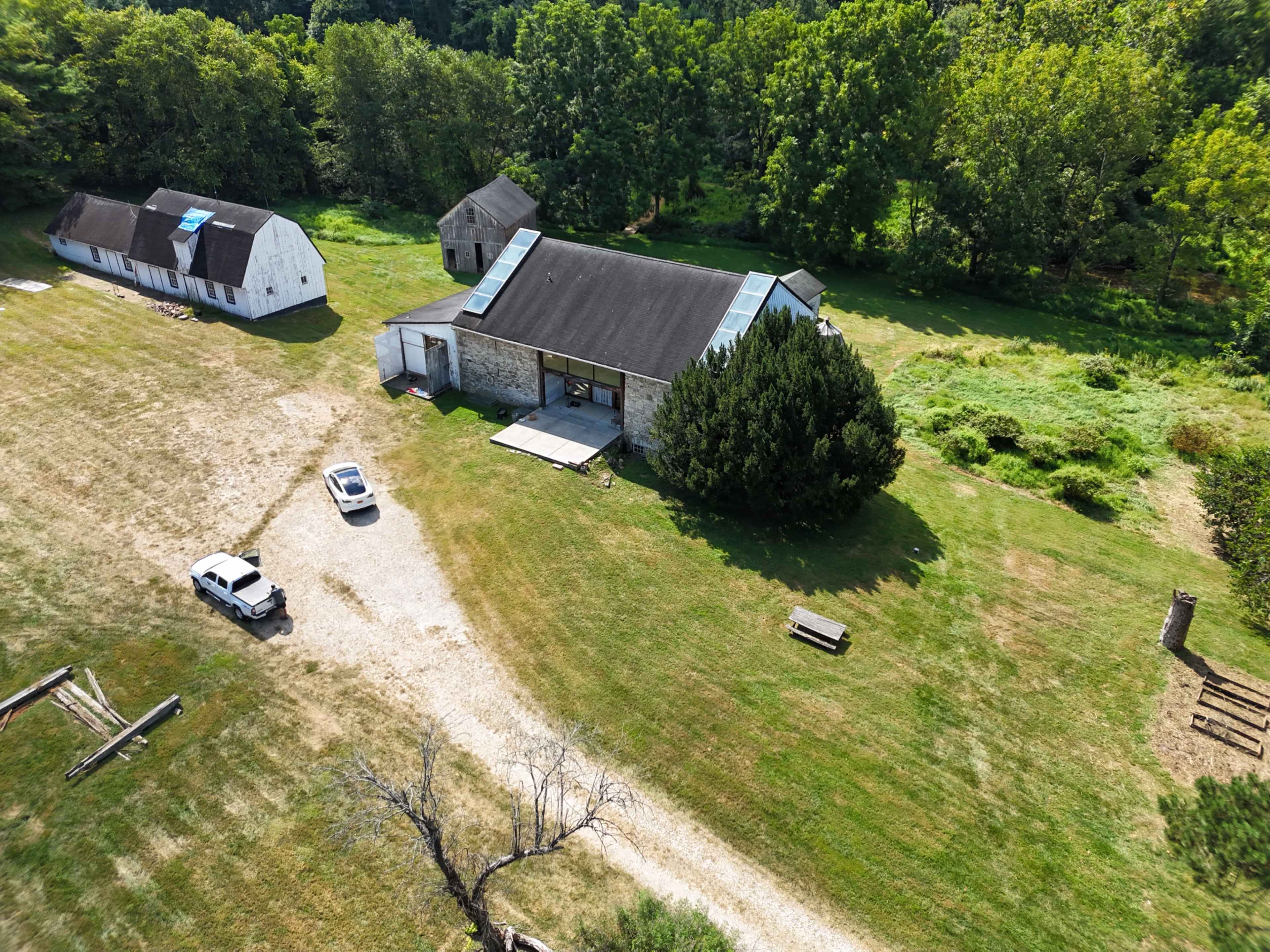 The image shows a rural landscape with a modern stone house surrounded by green grass, several trees, and a couple of outbuildings nearby.