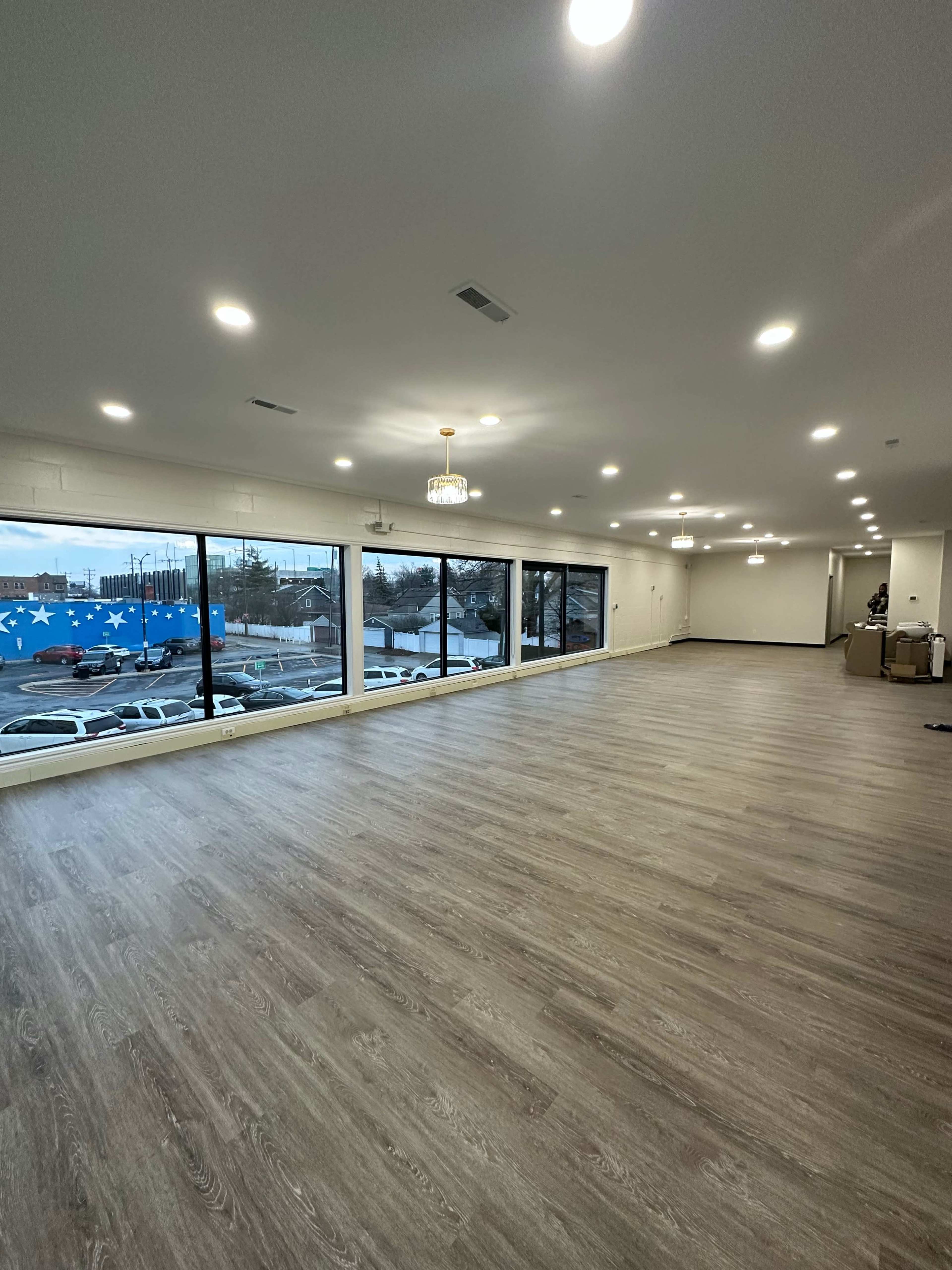 A spacious, empty room with wooden flooring and large windows overlooking a parking lot.