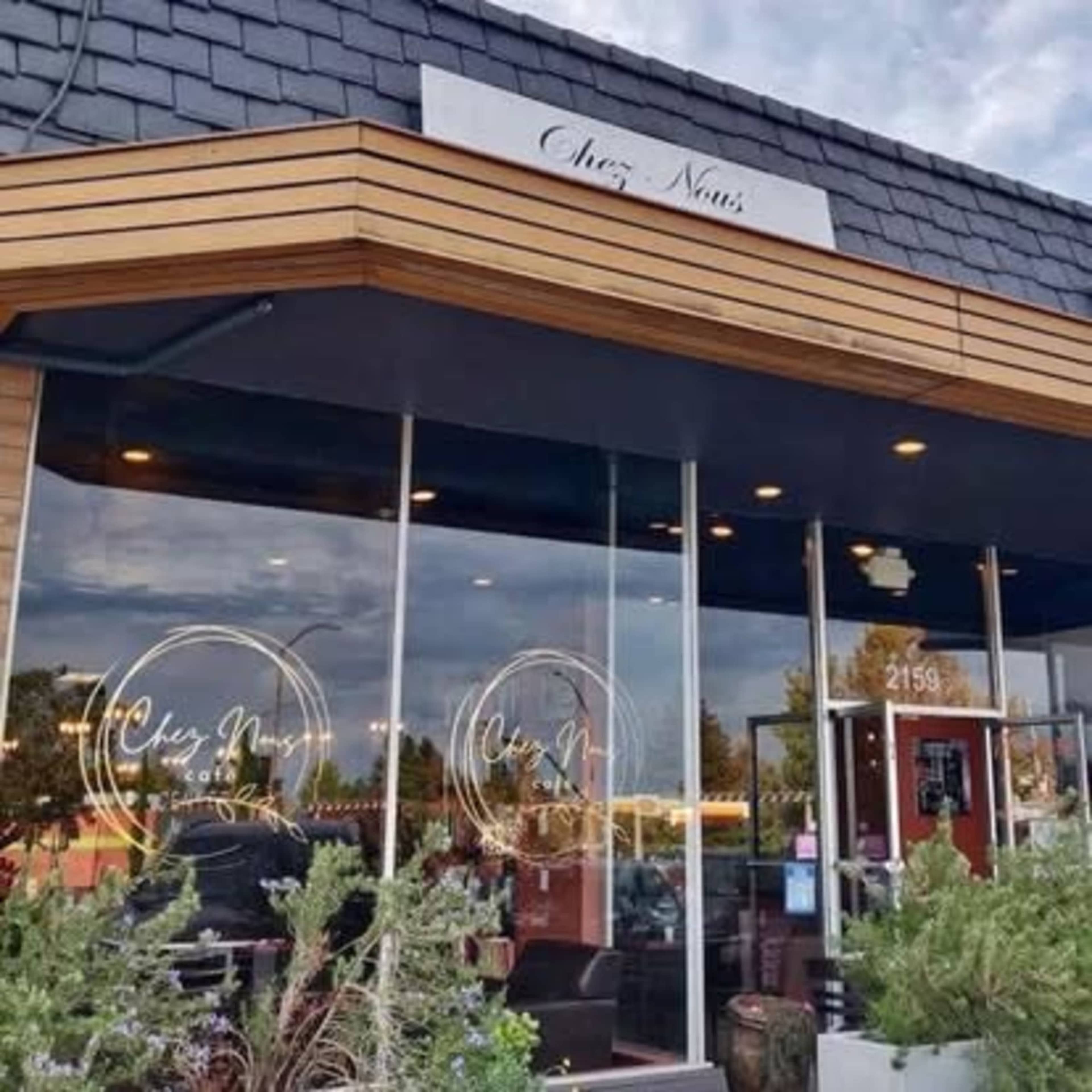 The image shows the exterior of a restaurant named "Chez Nous," featuring large glass windows and a modern wooden façade.