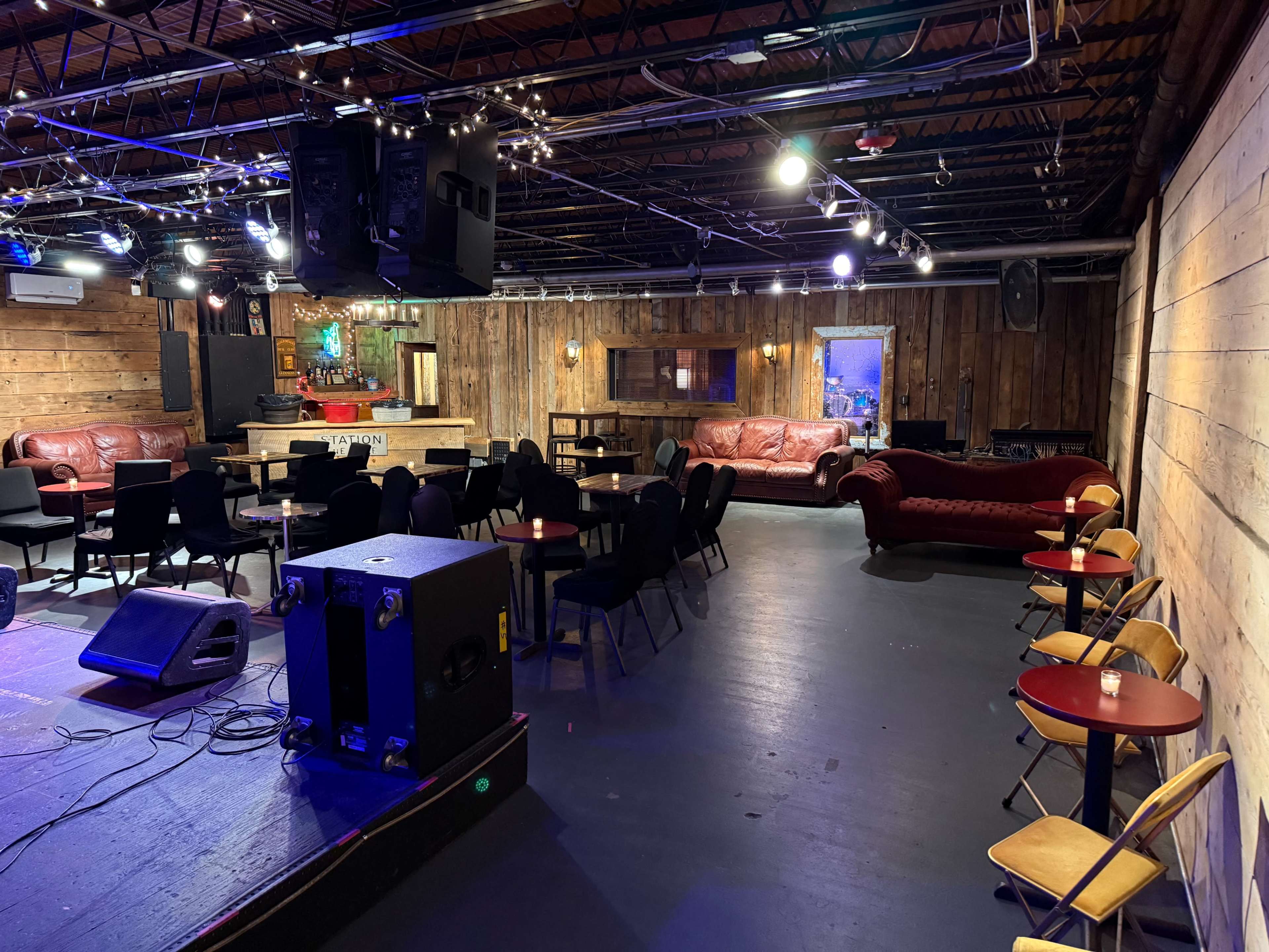 The image shows a dimly lit entertainment venue with wooden walls, seating areas including tables and chairs, and a stage with speakers at the front.
