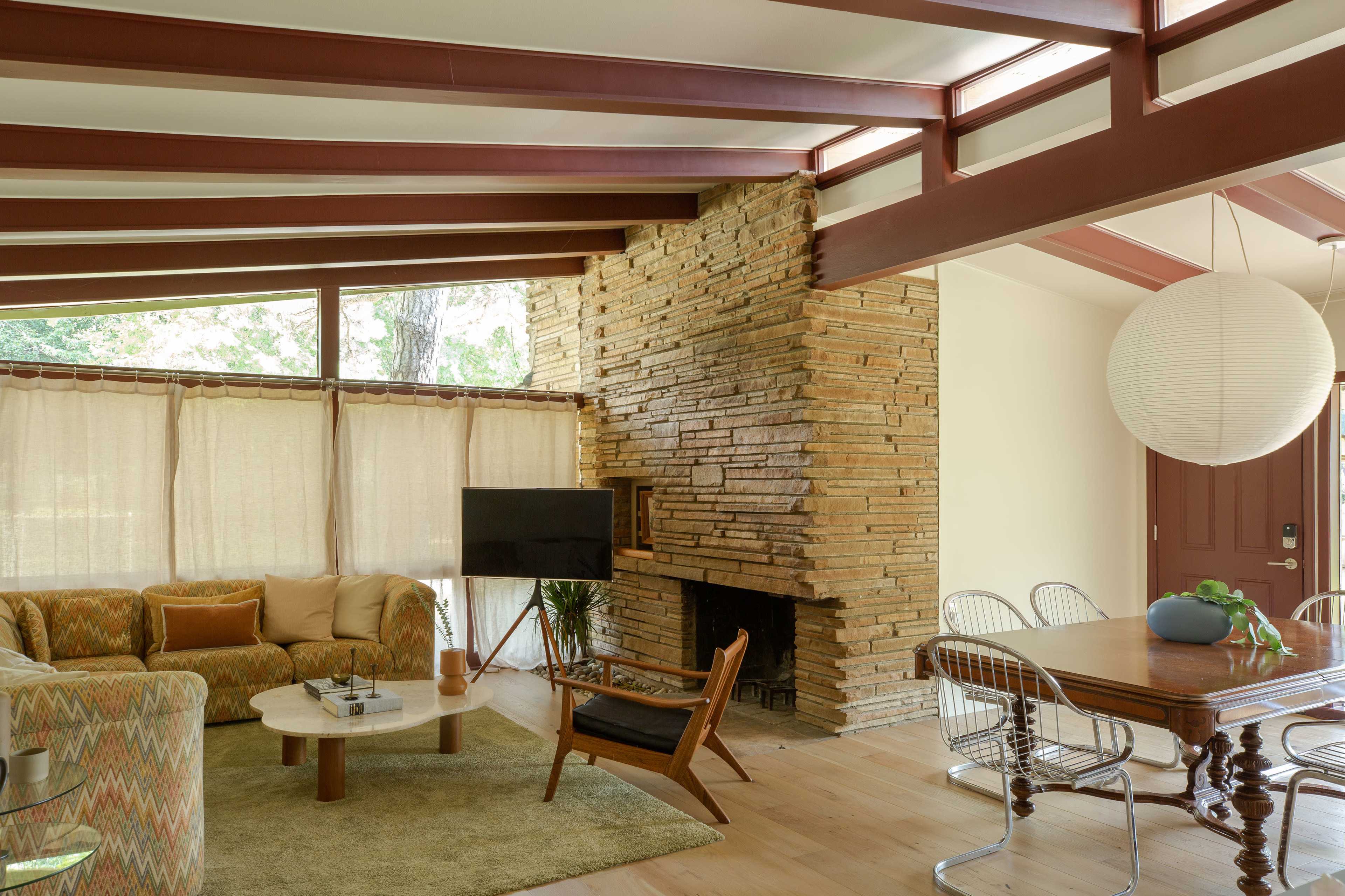 The image shows a mid-century modern living room featuring a stone fireplace, a circular coffee table, a beige sofa, and a dining area with a wooden table and metal chairs.