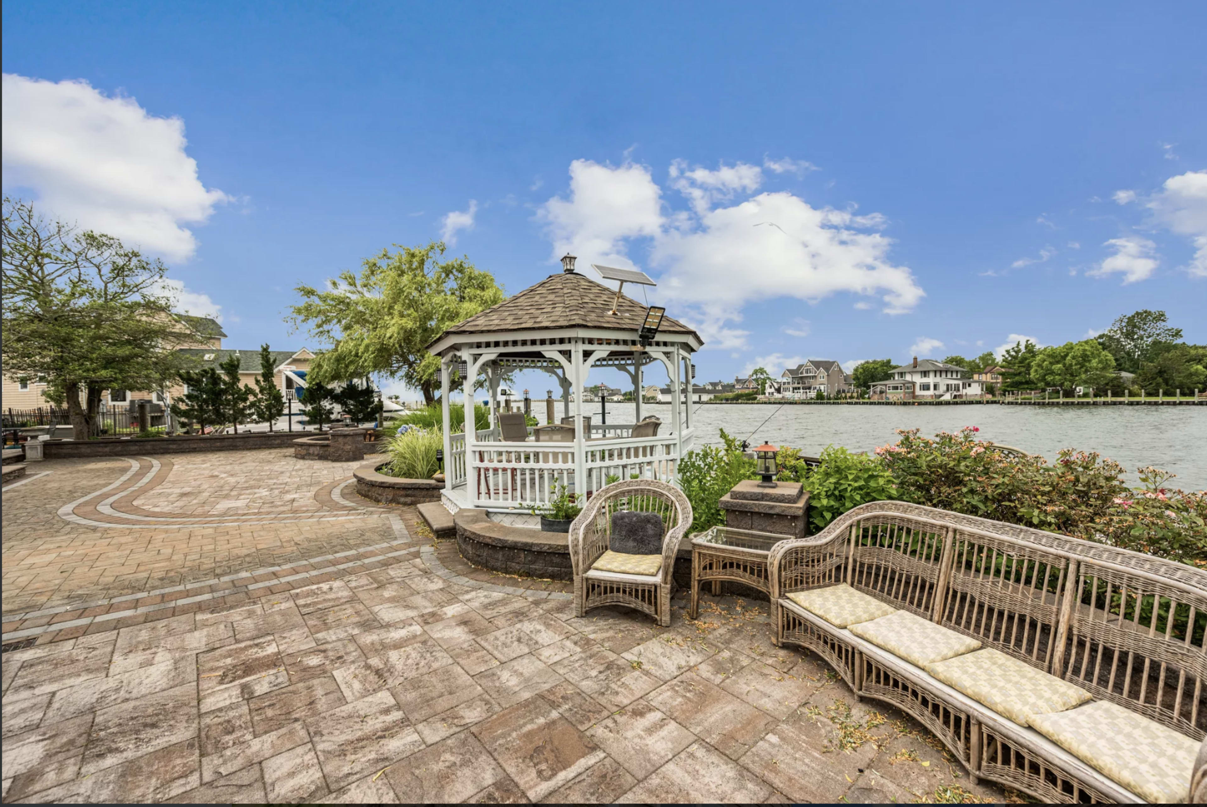 A gazebo stands by a waterfront, surrounded by a brick patio and wicker furniture.