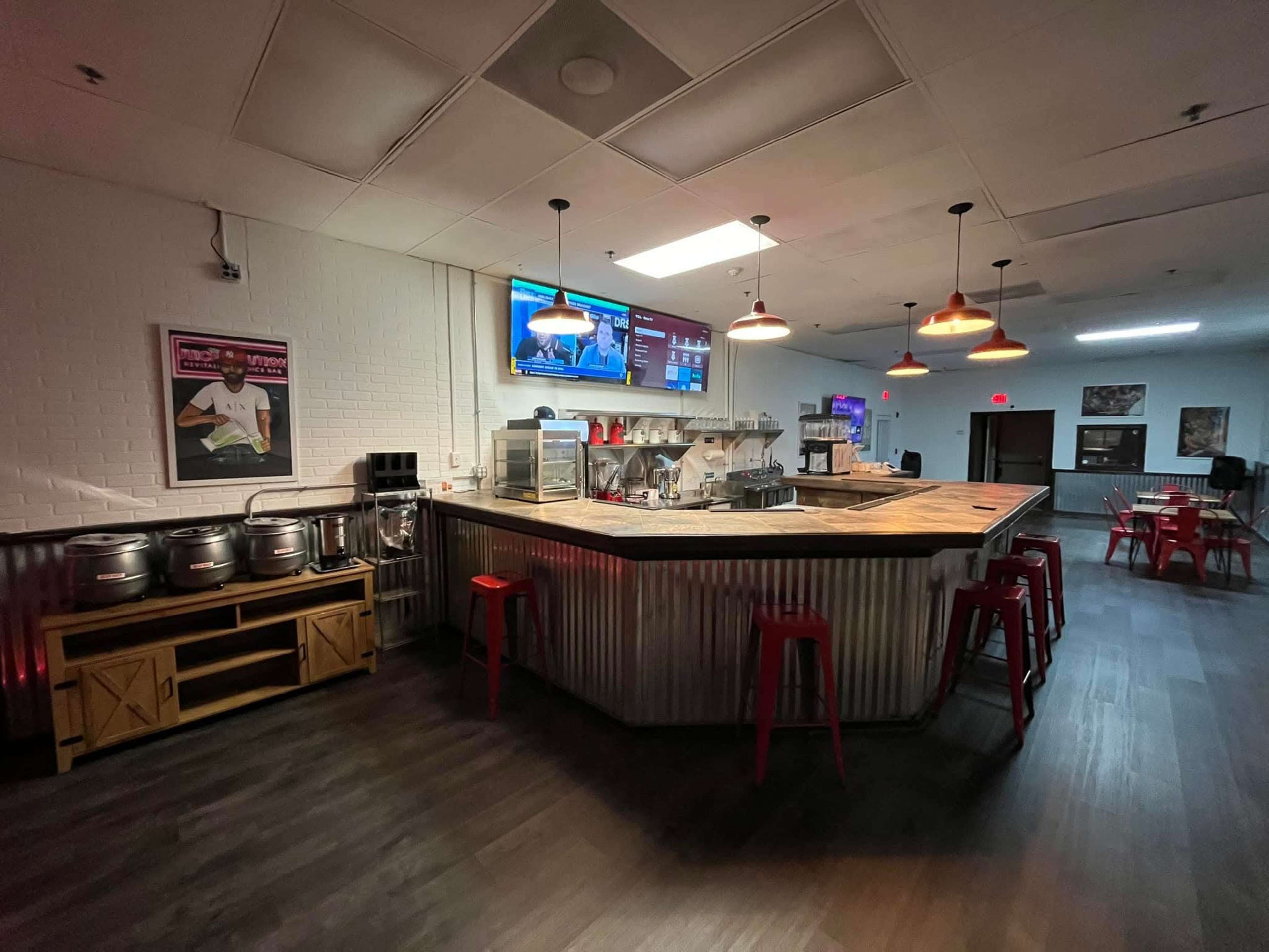 The image shows a modern bar area with a wooden counter, metal accents, and several red stools, along with multiple beverage dispensers and a television on the wall.