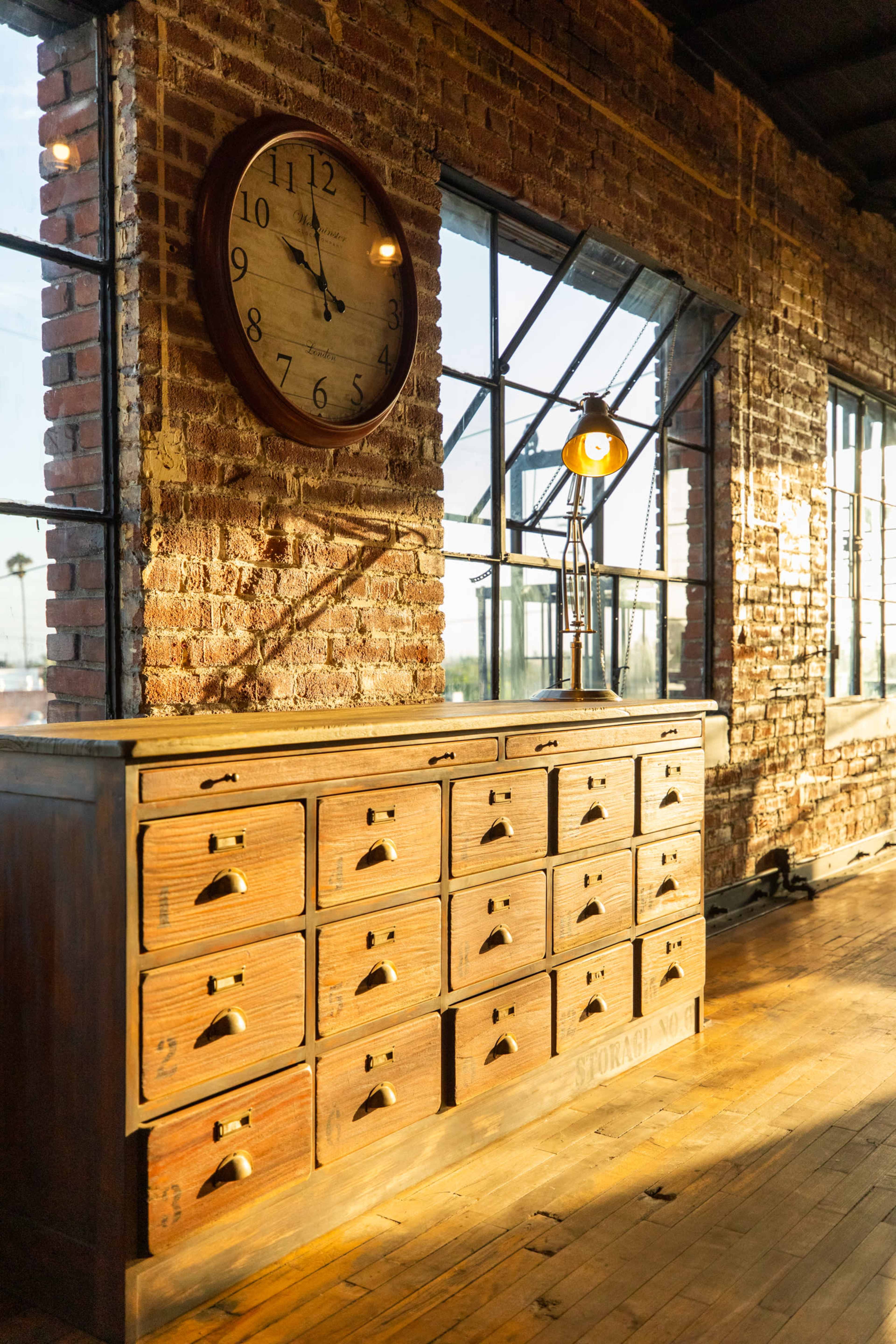 A wooden storage cabinet with multiple drawers, a wall clock, and large windows in a room with exposed brick walls.