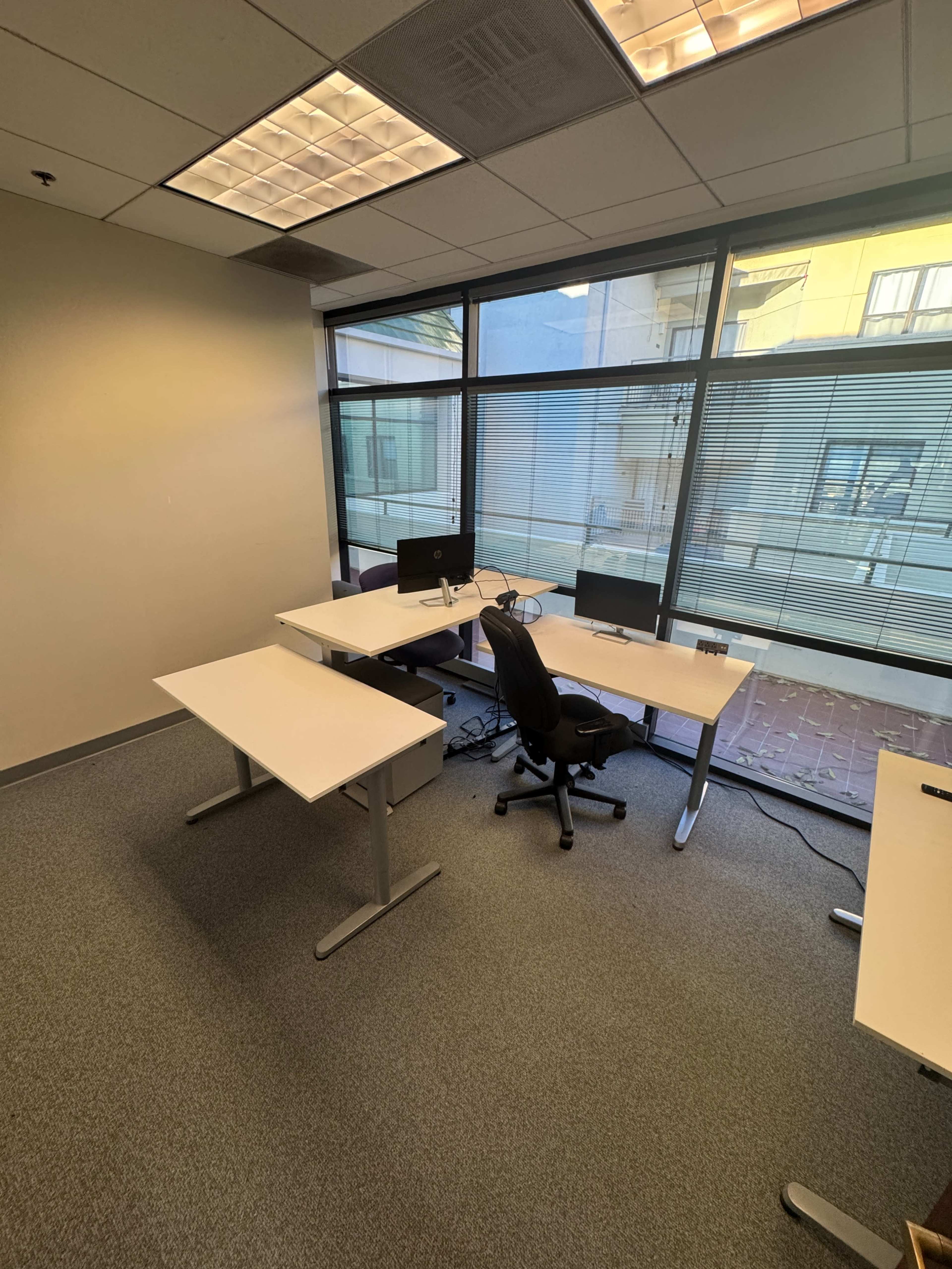 The image shows an office space with two white desks and a black office chair, located near large windows that provide natural light.