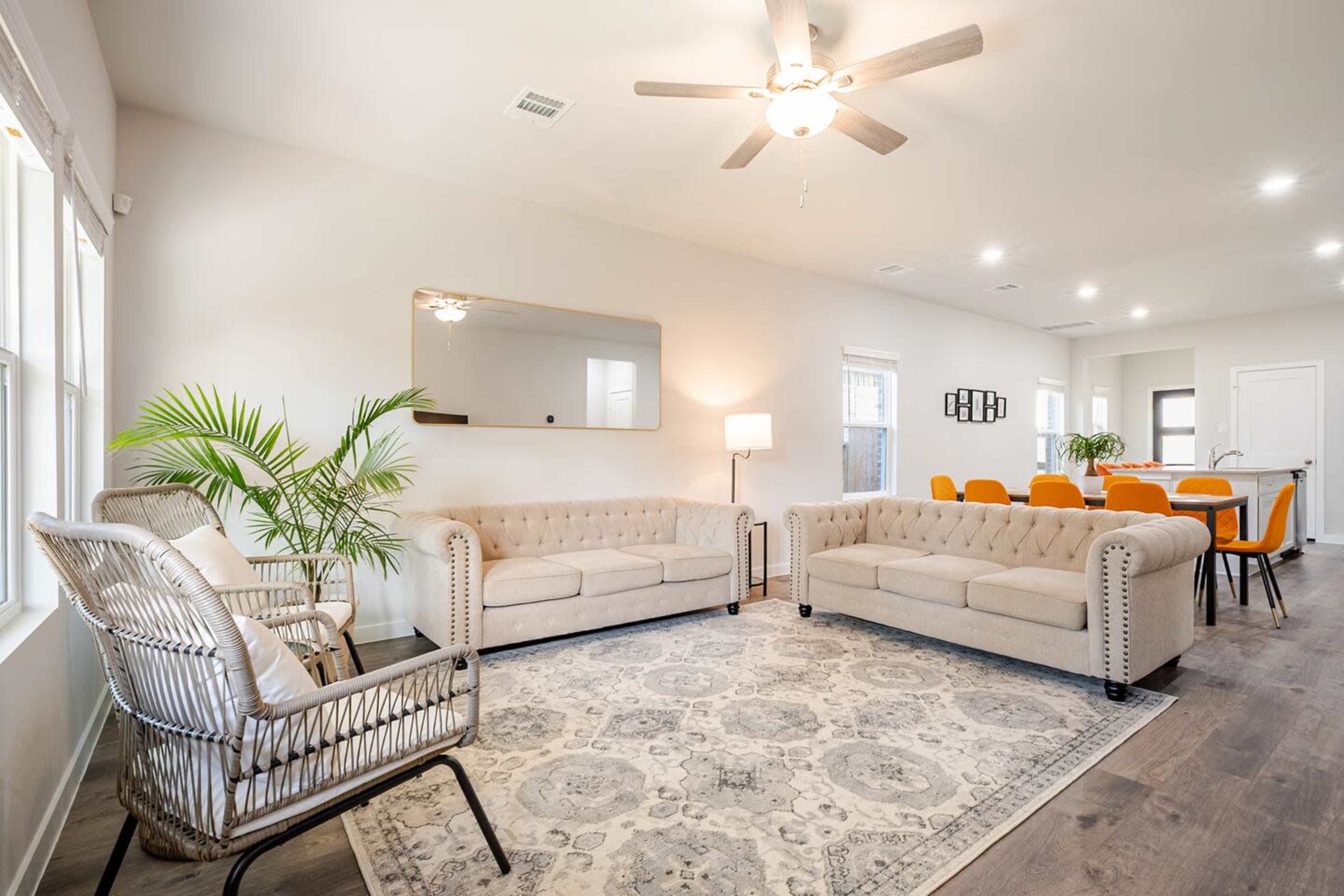 The image shows a modern living room with two tufted sofas, a large mirror on the wall, and a dining area in the background with orange chairs.