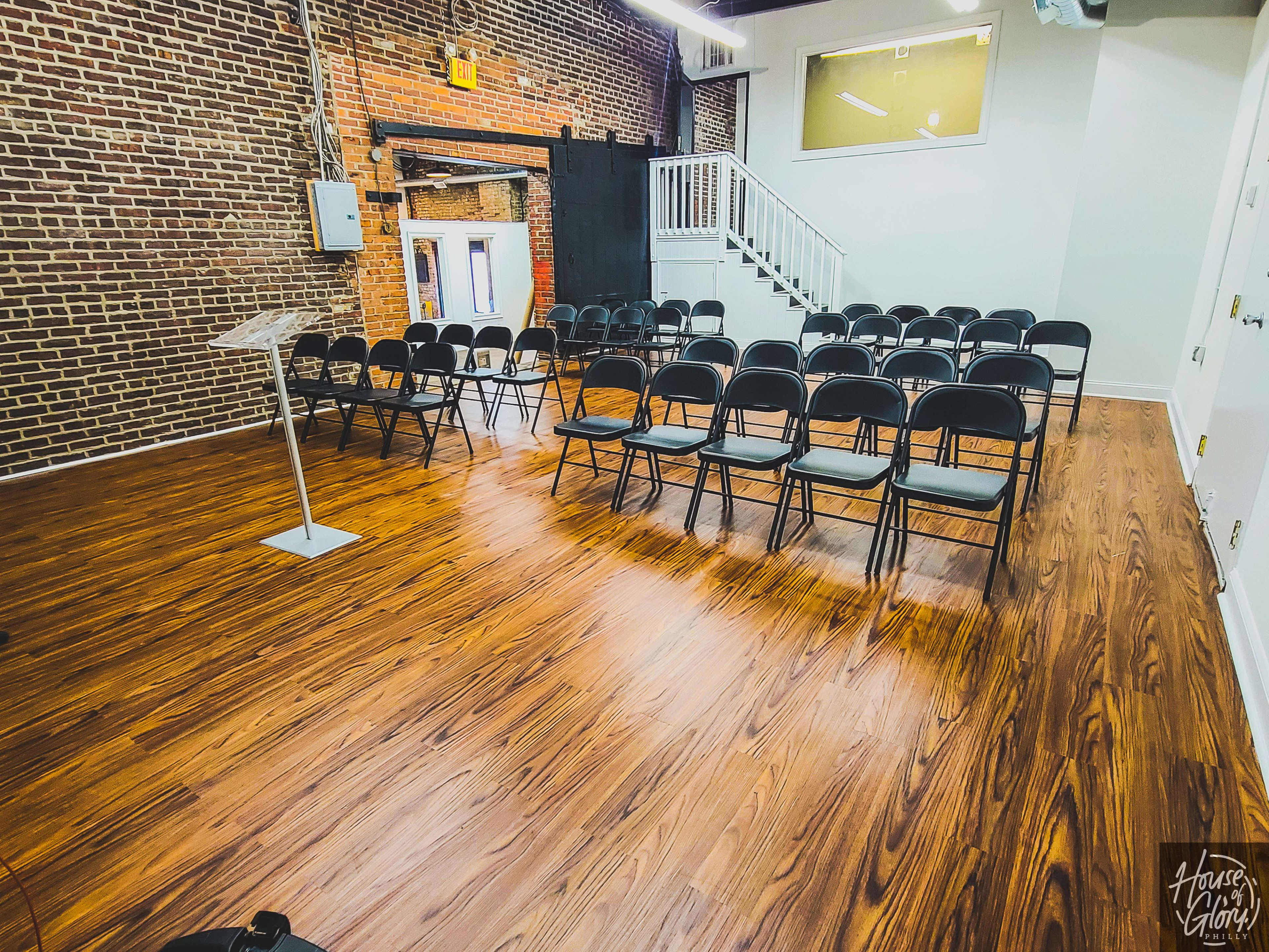 A room with a wooden floor features rows of black chairs facing a clear lectern, against a backdrop of exposed brick walls.