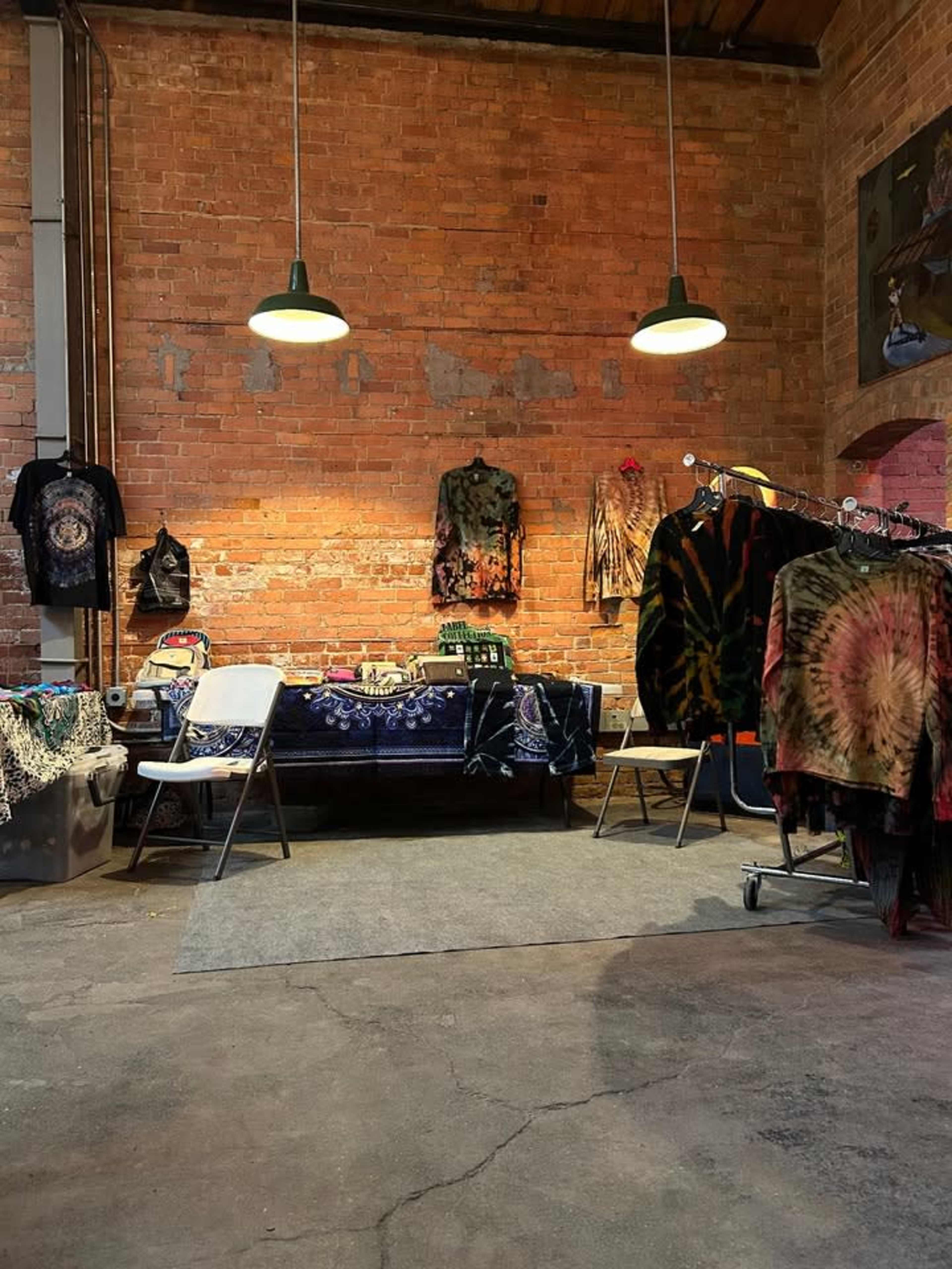 The interior of a rustic shop features exposed brick walls with various tie-dye clothing hanging on display and a table cluttered with merchandise beneath two hanging lights.