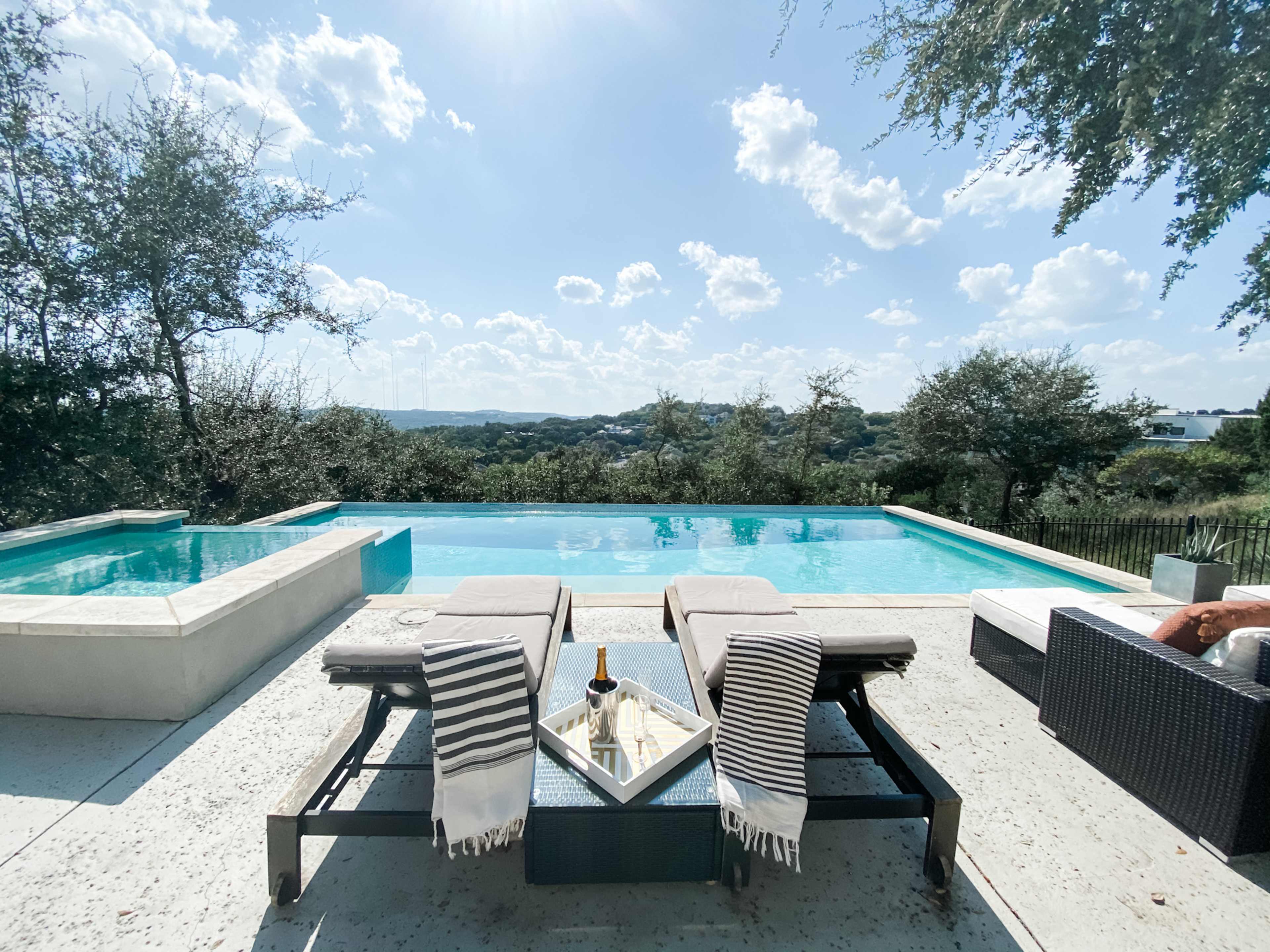 A clear swimming pool with lounge chairs and a table is set against a backdrop of rolling hills and a blue sky.