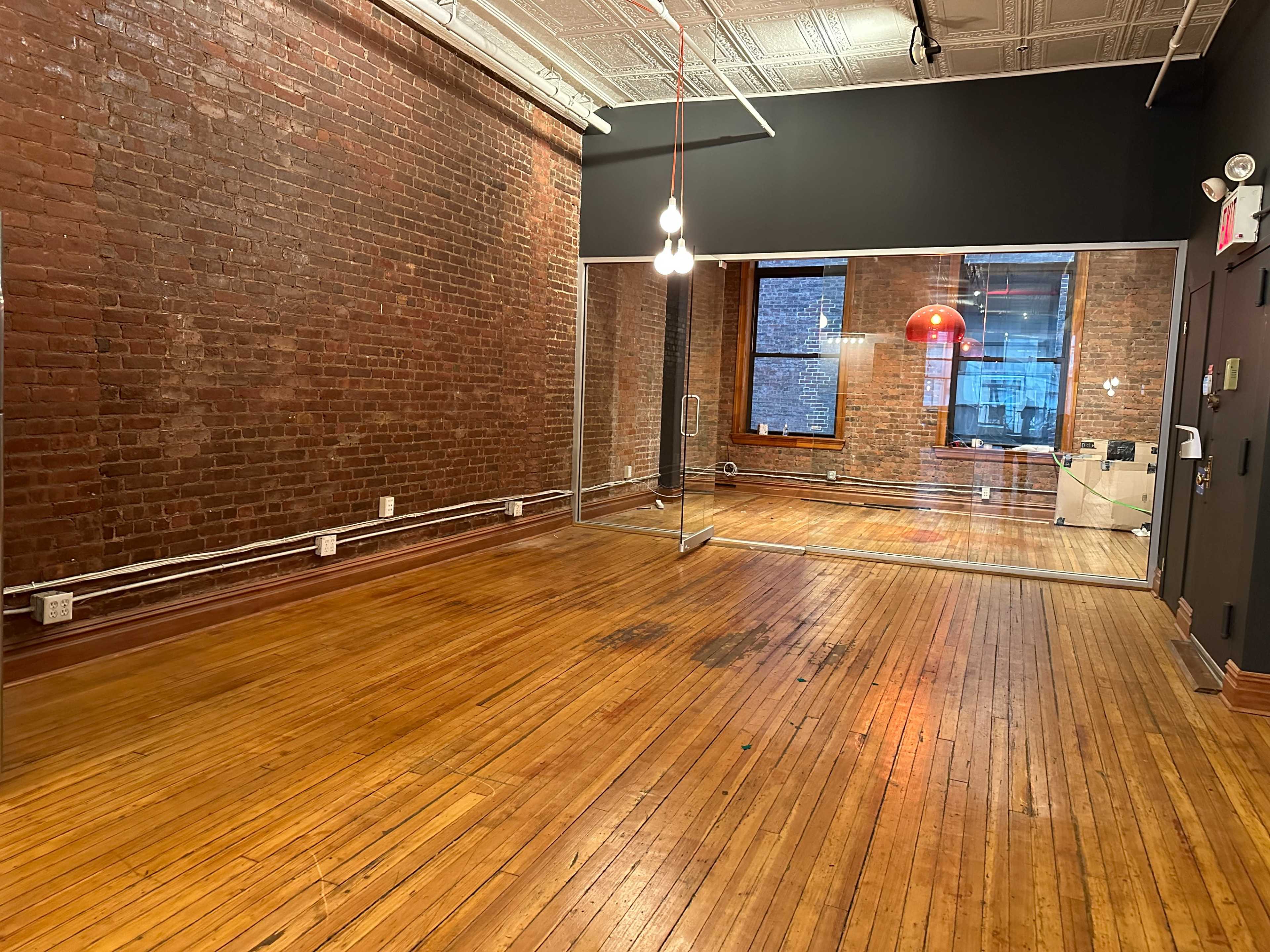 A spacious room with exposed brick walls, wooden flooring, and a large glass wall separating a smaller room with a red pendant light.