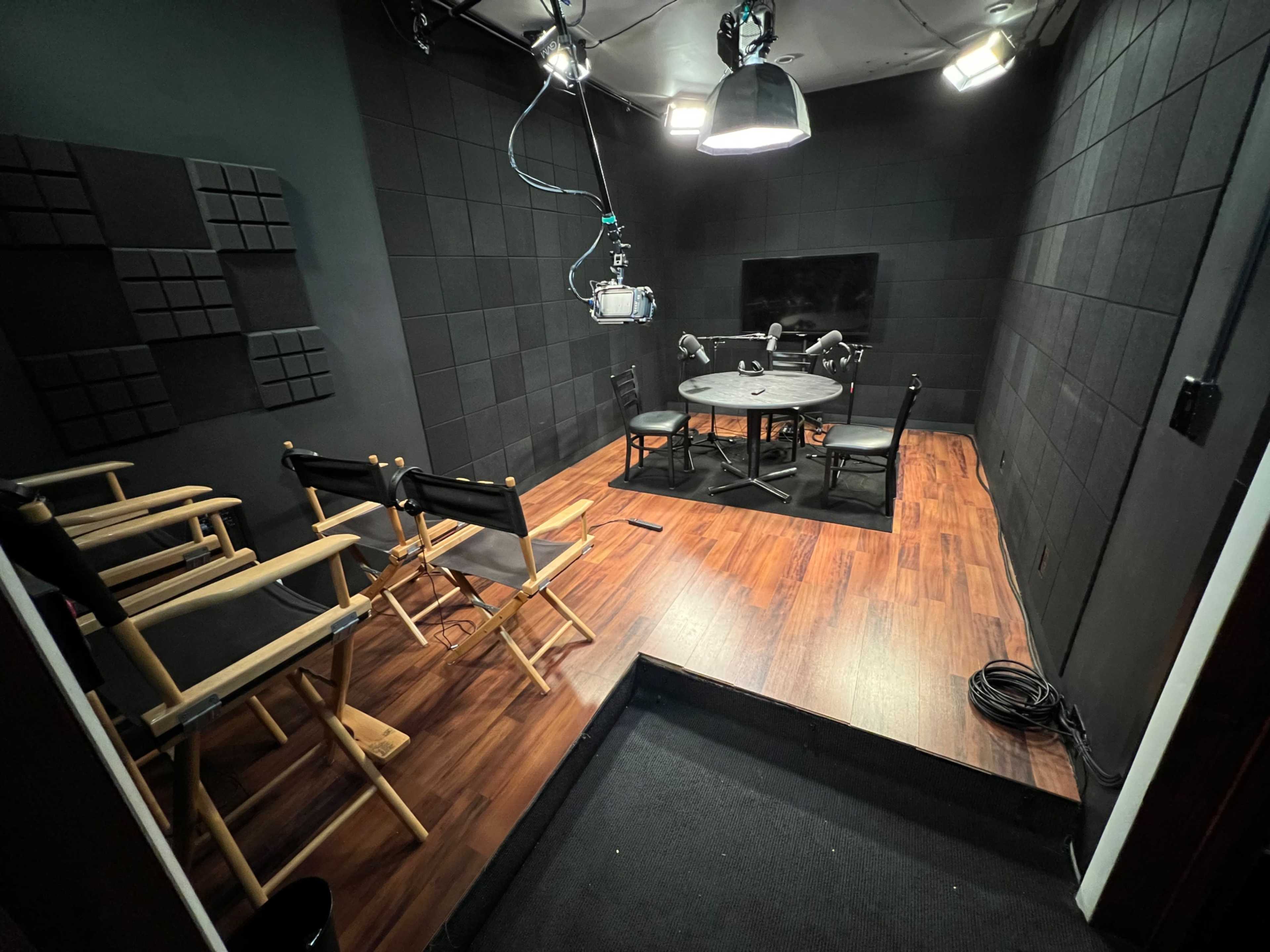 A dimly lit studio featuring a round table surrounded by chairs, with soundproofed walls and overhead lighting.
