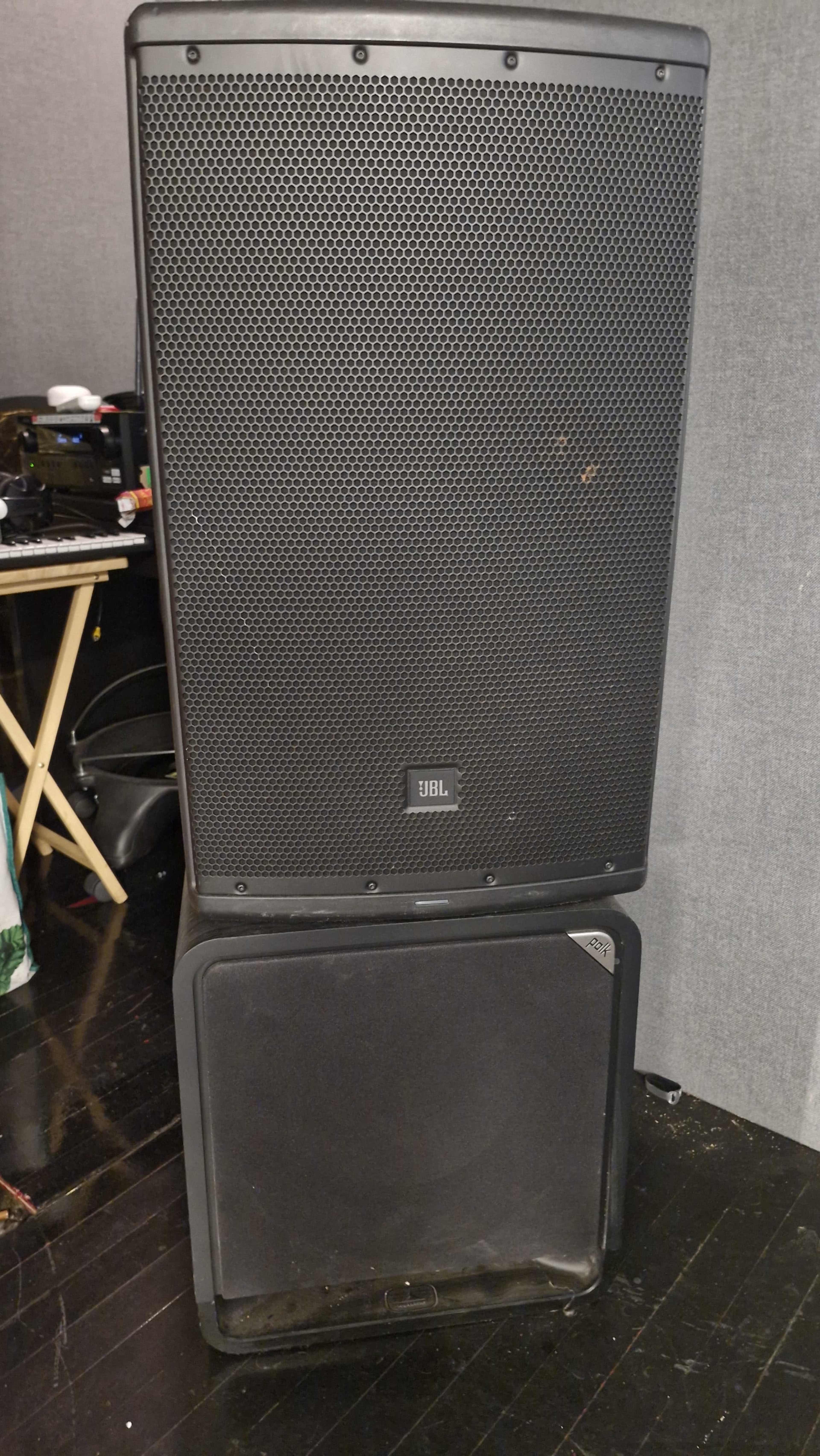The image shows a large black JBL speaker stacked on top of a black subwoofer in a dimly lit room.