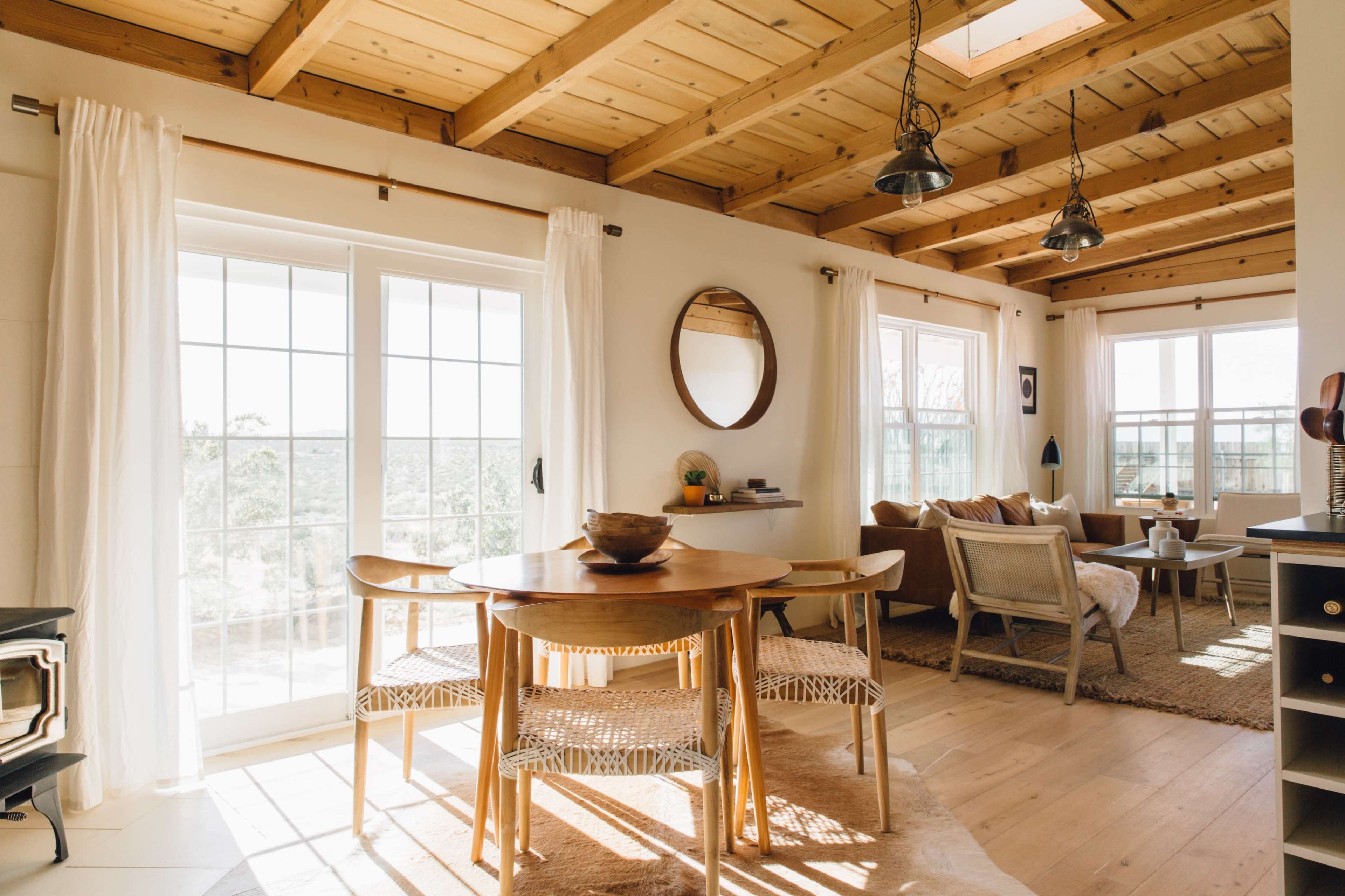 A cozy open-plan living space features a dining table near large windows, wooden beams overhead, and a comfortable seating area.