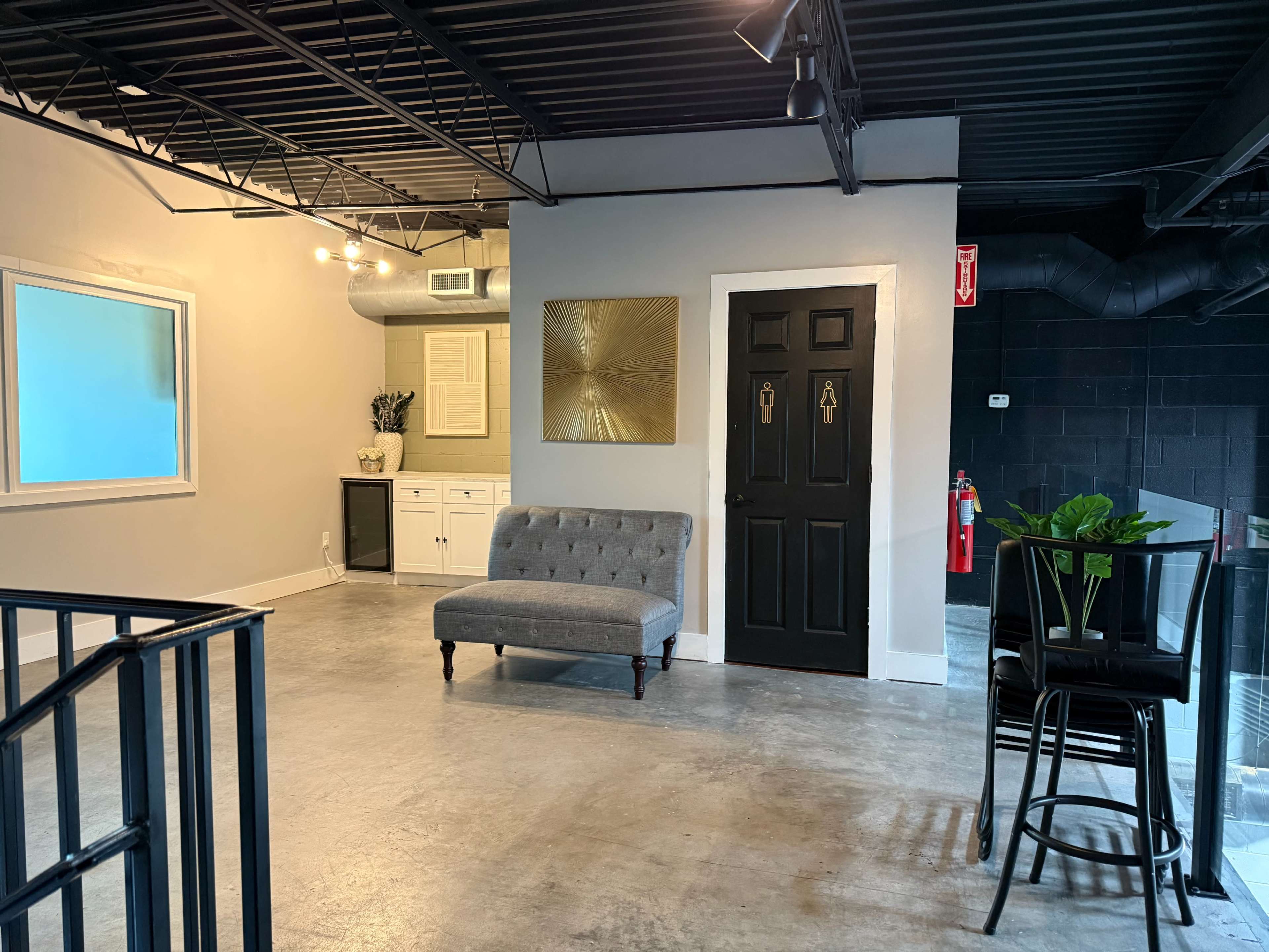 The image shows a modern, open interior space featuring a gray tufted sofa, a black door, and a bar stool near a glass partition.