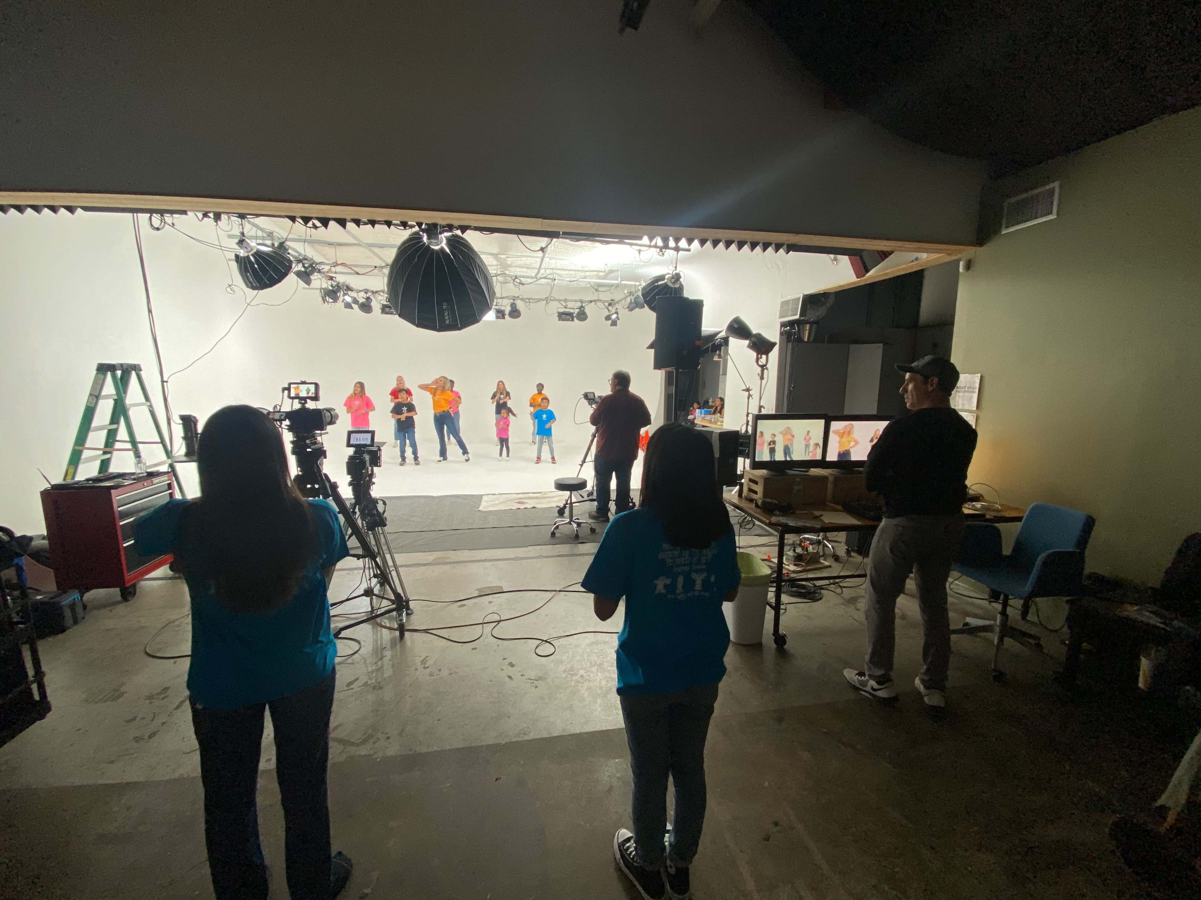 A group of performers is rehearsing in a studio while a few people operate cameras and equipment from the back.
