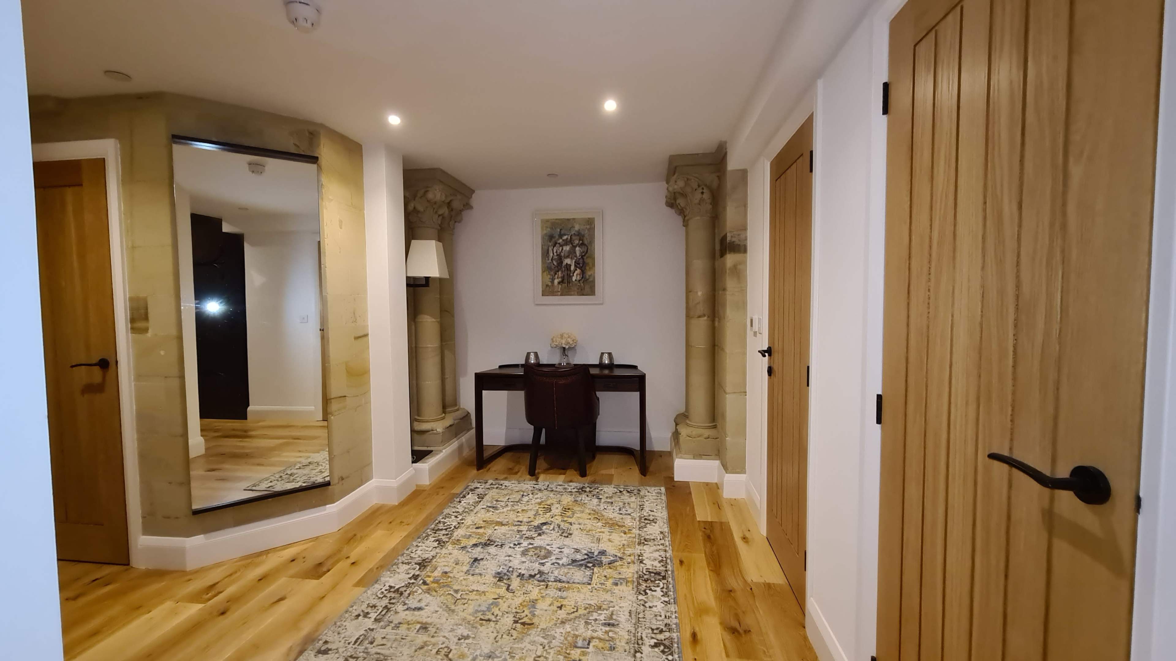 A hallway featuring a large mirror, a wooden desk with a chair, and stone columns alongside a decorative rug on a wooden floor.