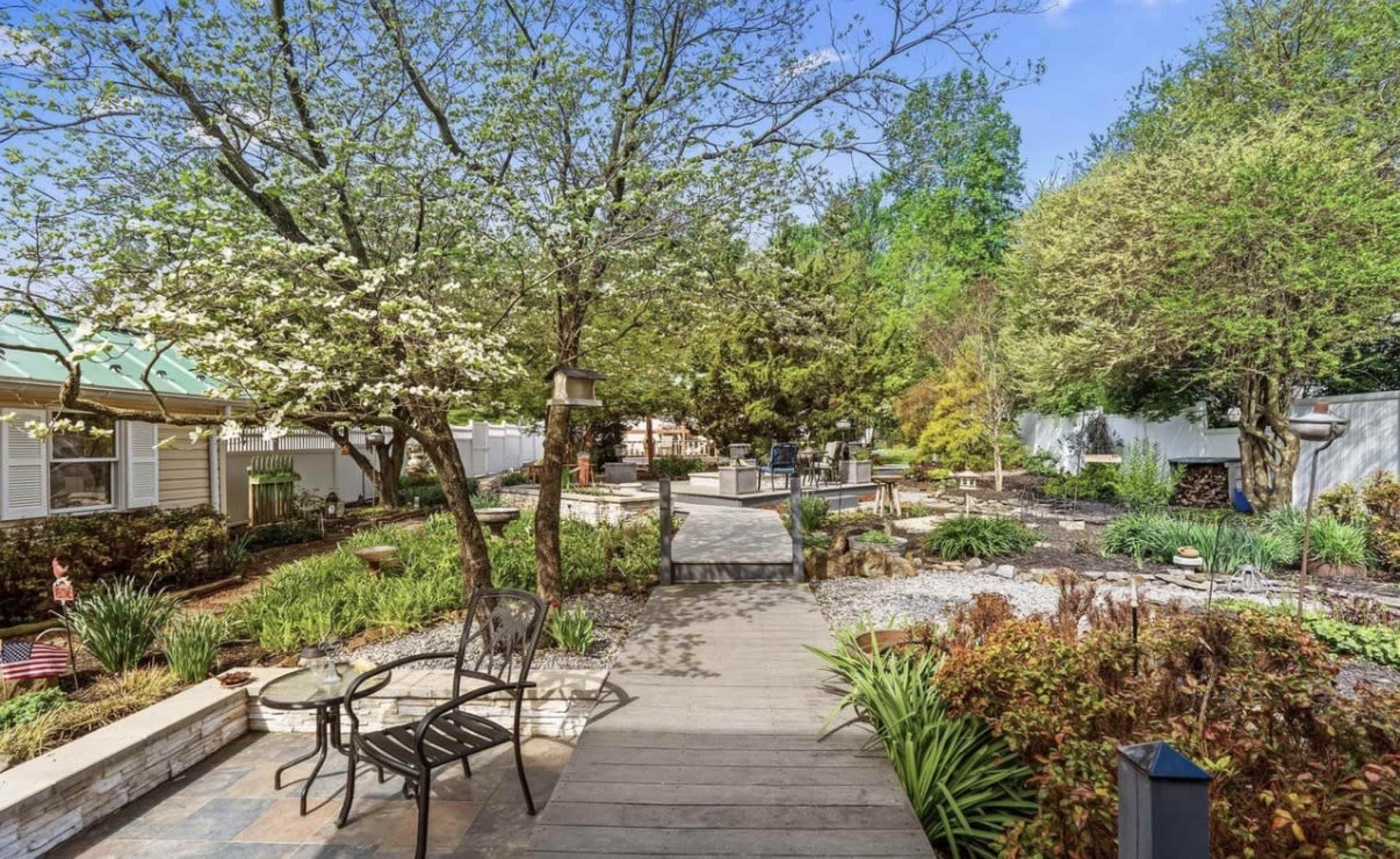 A wooden pathway leads through a landscaped garden with various plants, trees, and seating areas.