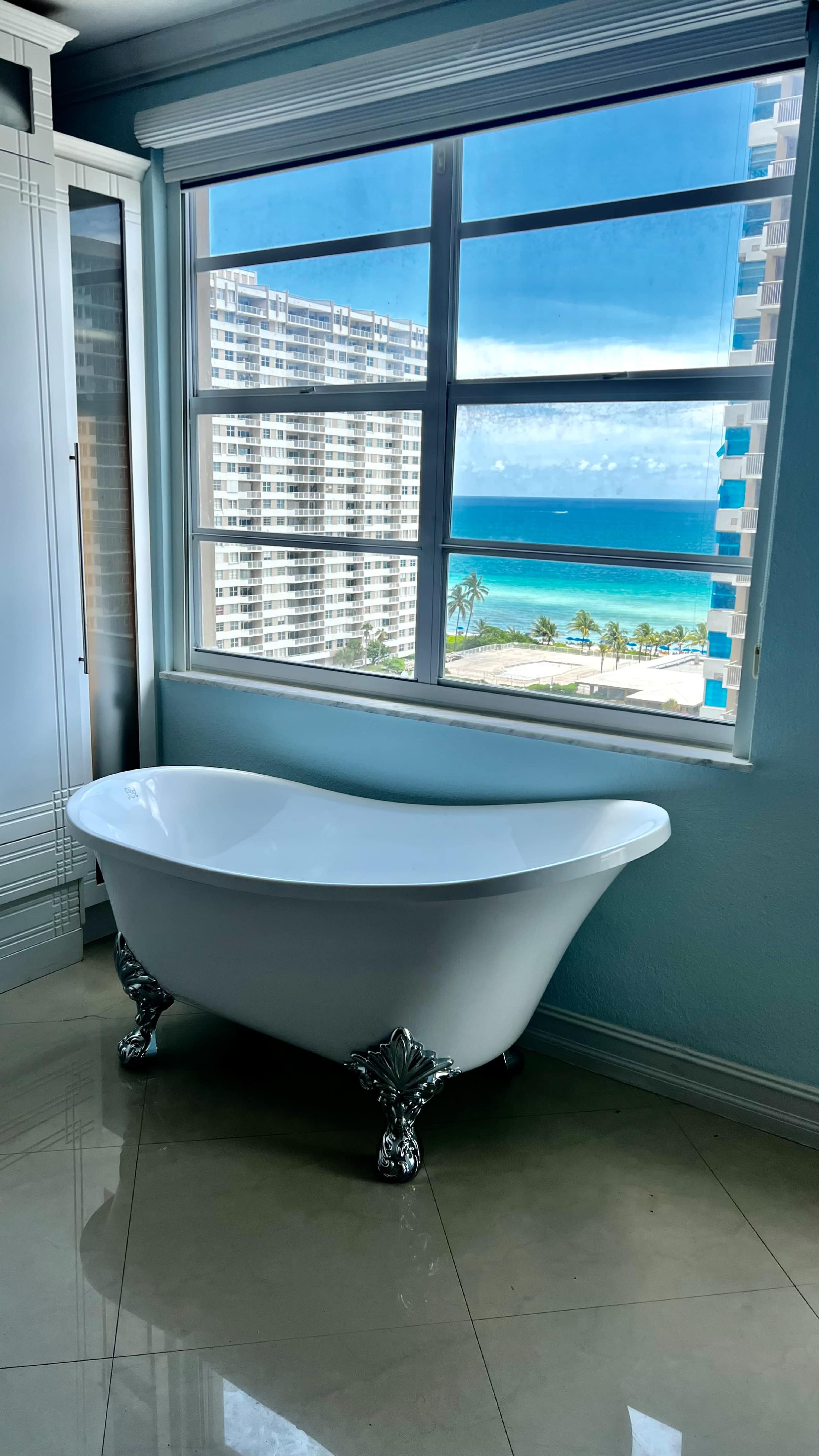 A freestanding white bathtub with chrome feet is positioned near a large window overlooking a view of the ocean and buildings.