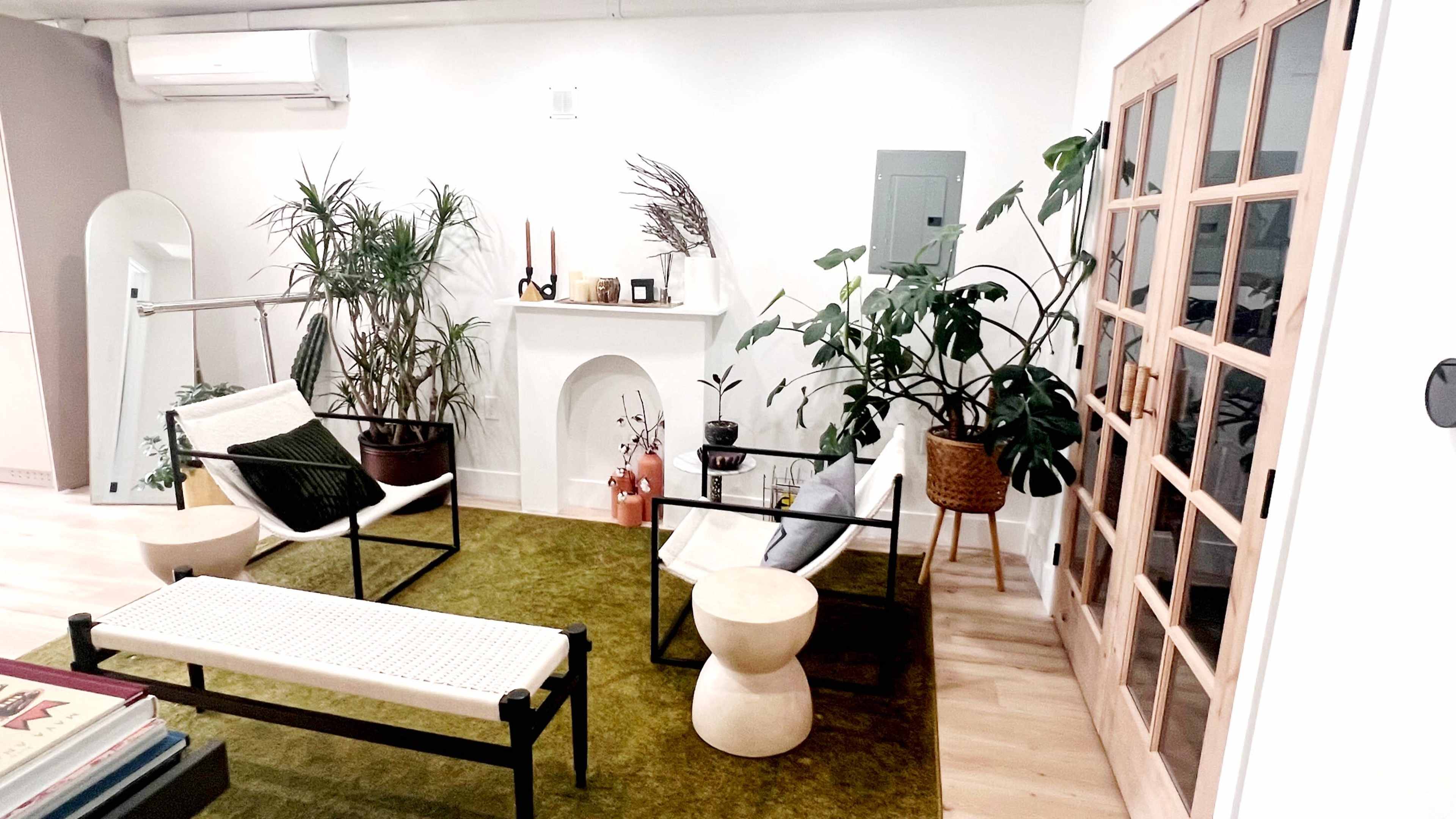 A modern interior setting with two lounge chairs, a small round table, a bench, various potted plants, and a decorative fireplace, all arranged on a green rug.