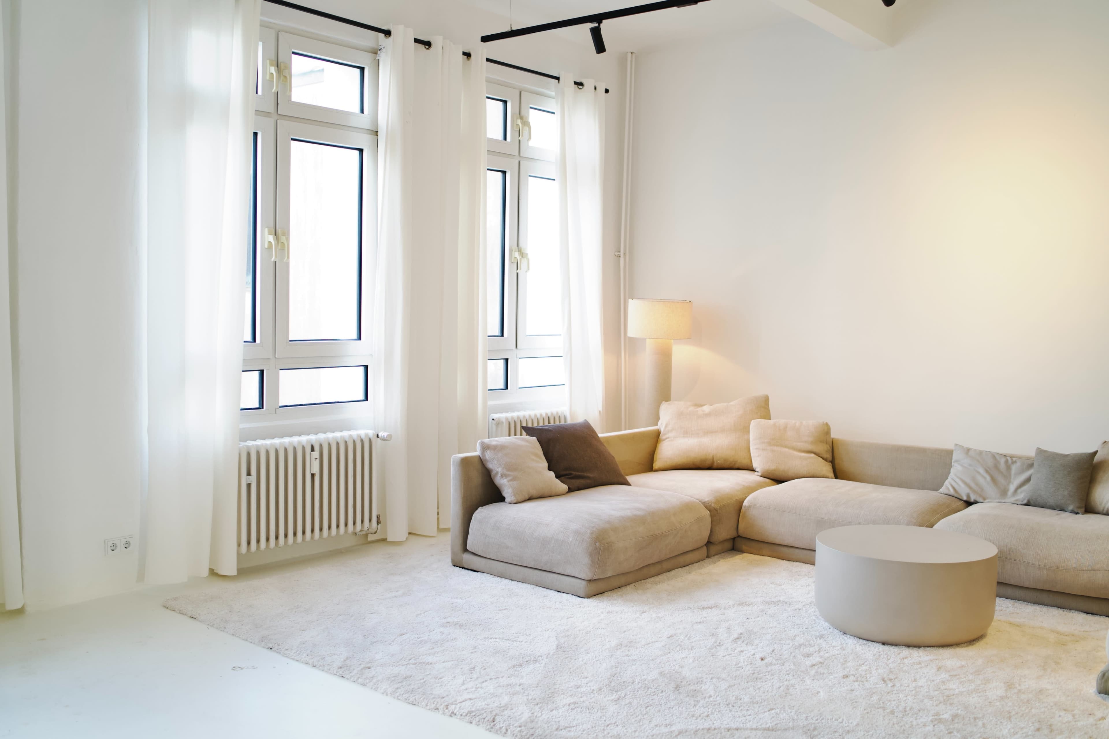 The image shows a partially furnished living room with a large sectional sofa, a round coffee table, and ample natural light from the windows.