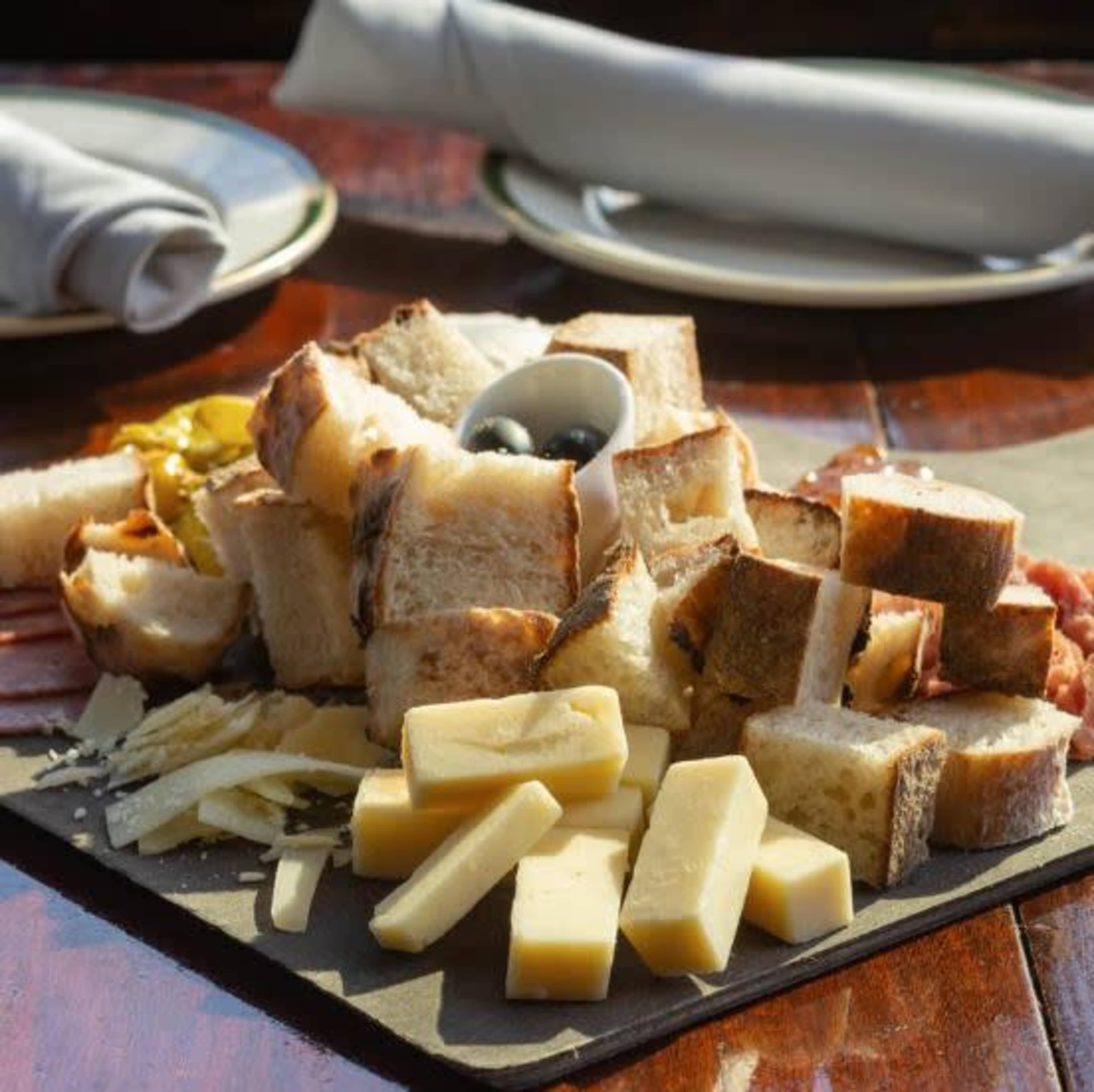 A selection of sliced bread and cheese is arranged on a wooden platter, accompanied by olives and other garnishes.