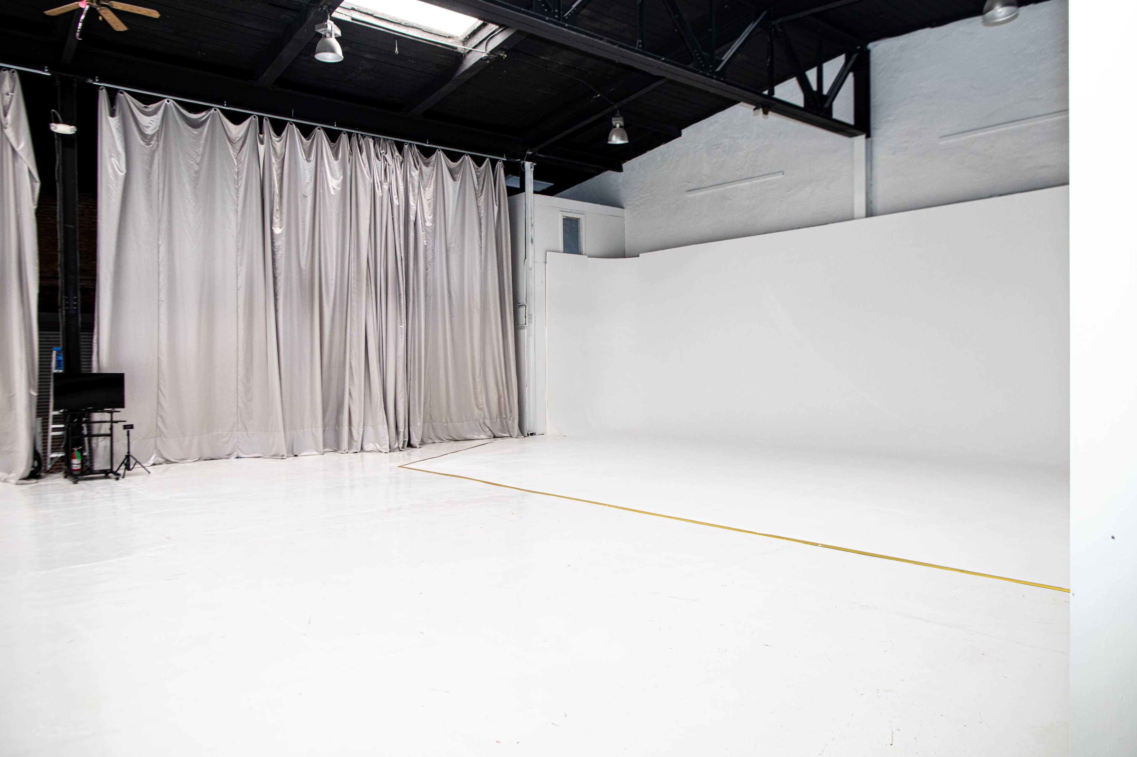The image shows a spacious, empty studio with white walls and a polished floor, featuring gray curtains on one side and a small table with a chair in the corner.