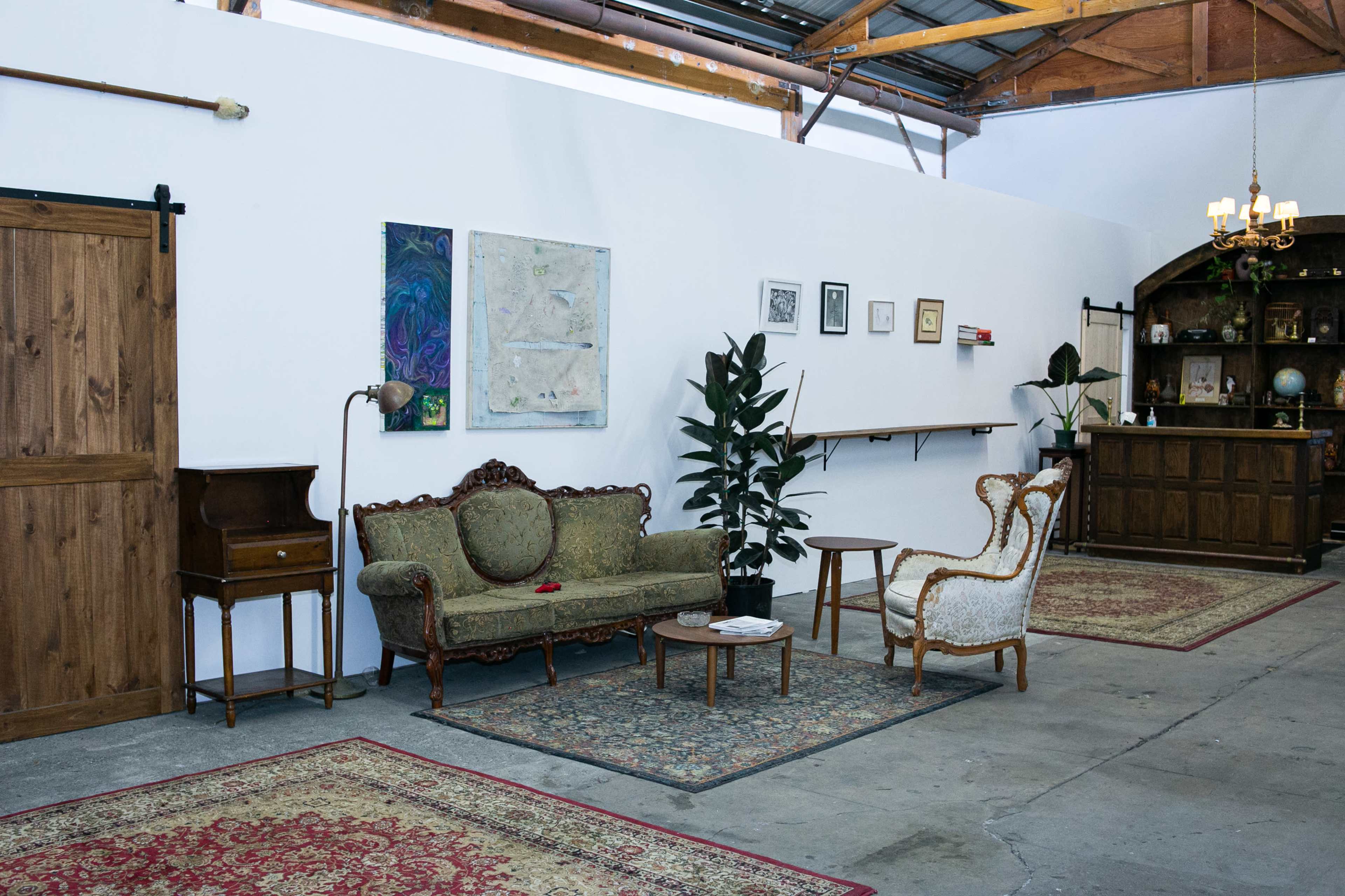The image shows a spacious room featuring a vintage sofa, a decorative chair, a small table, potted plants, and various framed artworks on the walls.