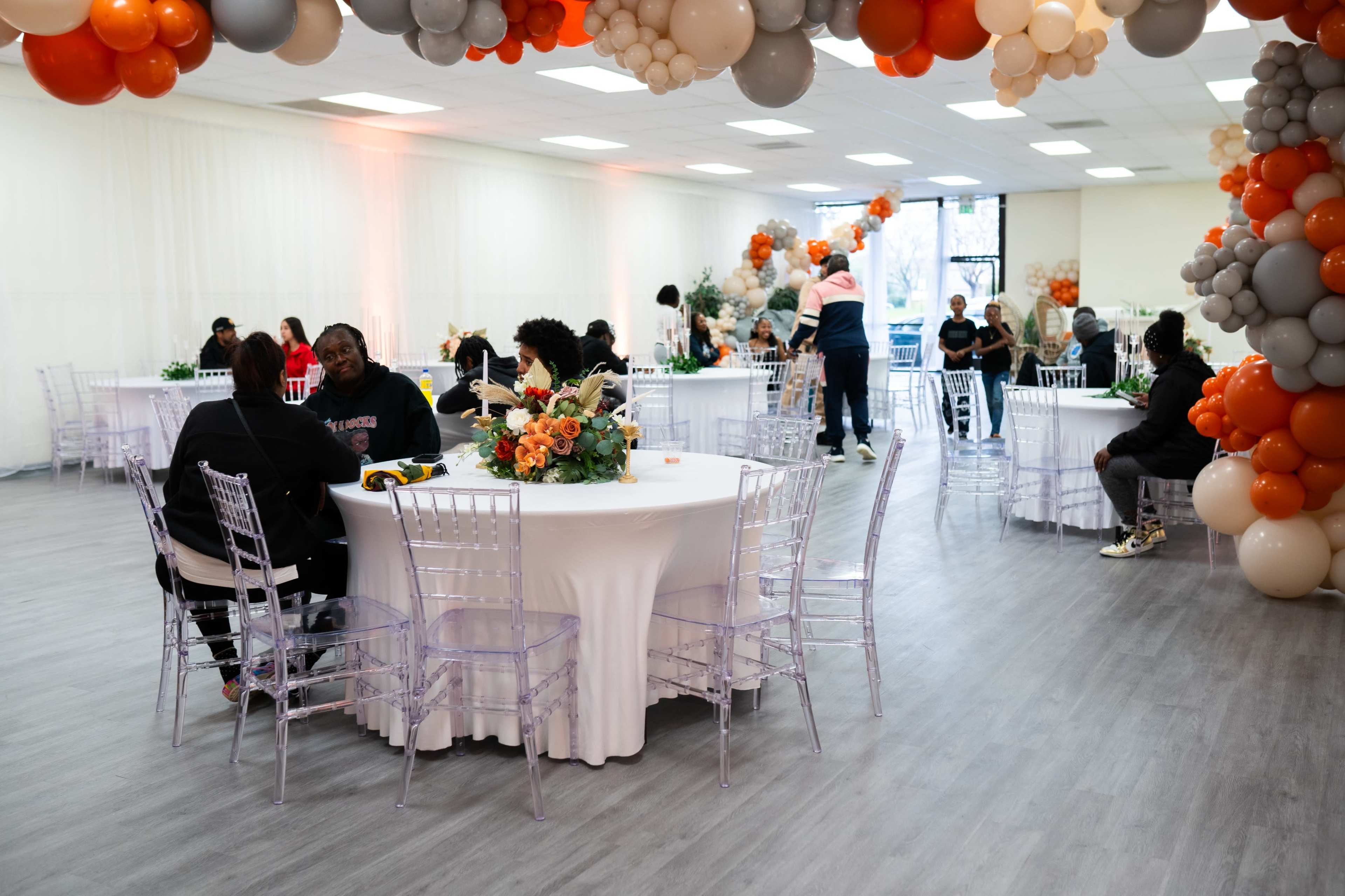 A spacious event hall is set up with round tables covered in white linens, clear chairs, and decorated with floral centerpieces, while balloons adorn the ceiling.