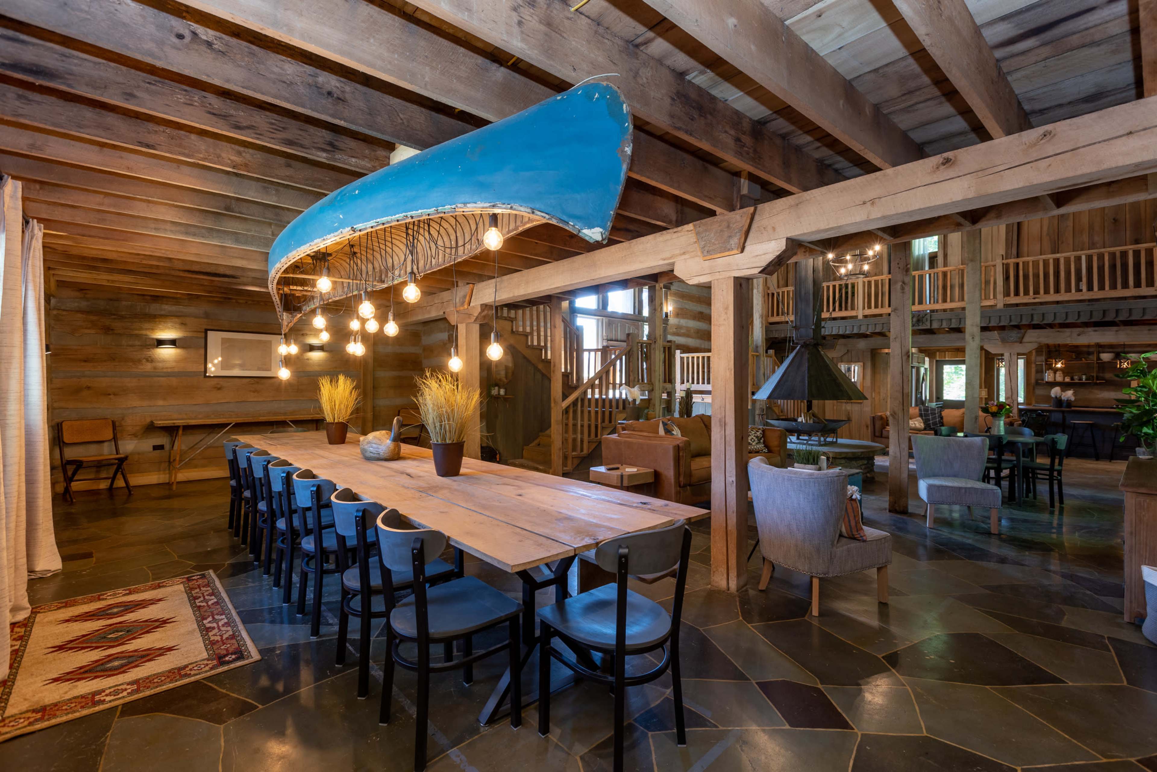 A long wooden dining table is set under a suspended blue canoe, illuminated by multiple hanging light bulbs in a spacious, rustic interior with wooden beams.