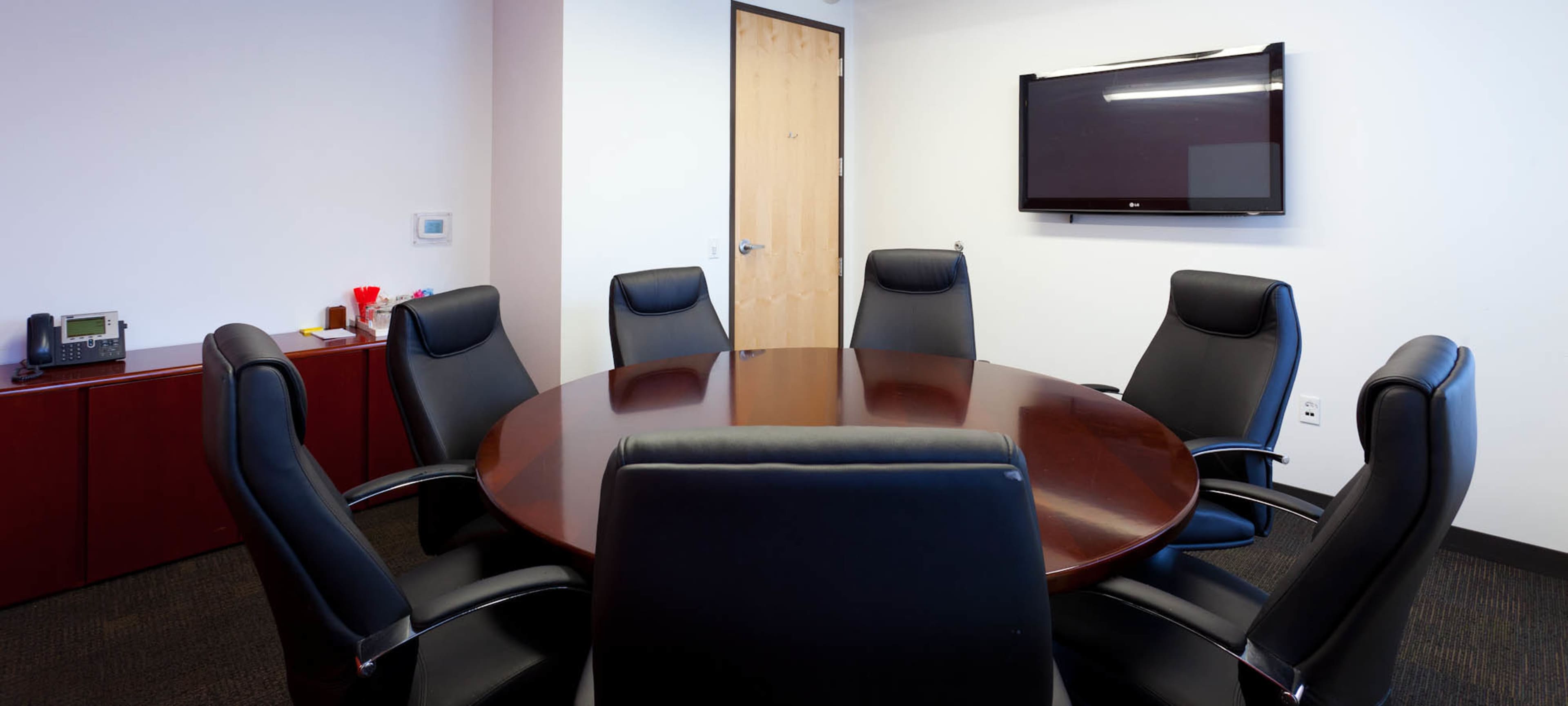 A circular wooden conference table is surrounded by six black leather chairs in a minimalist meeting room with a wall-mounted television and a wooden door.