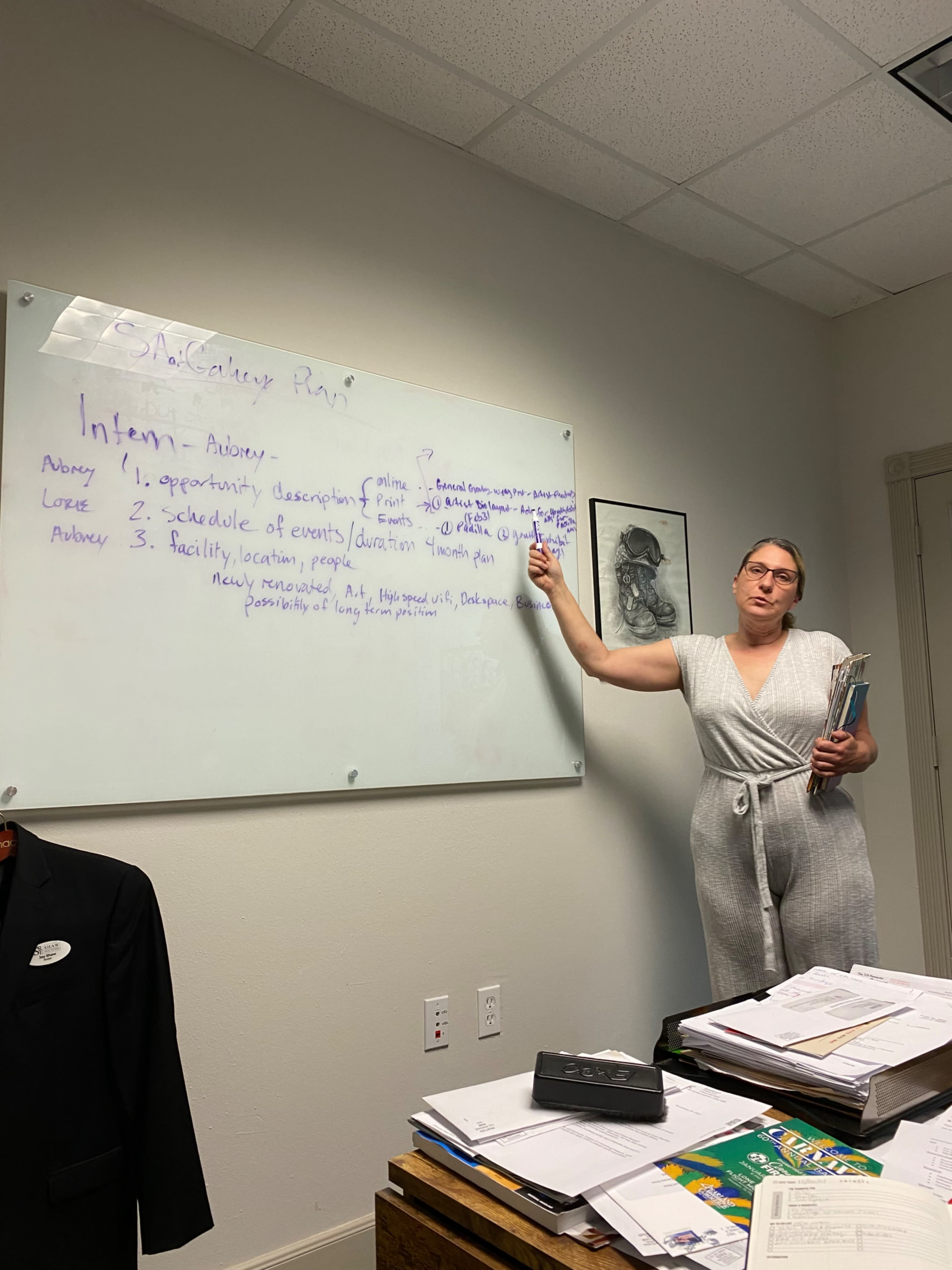 A woman in a white outfit points at a whiteboard covered with handwritten notes in an office cluttered with papers.