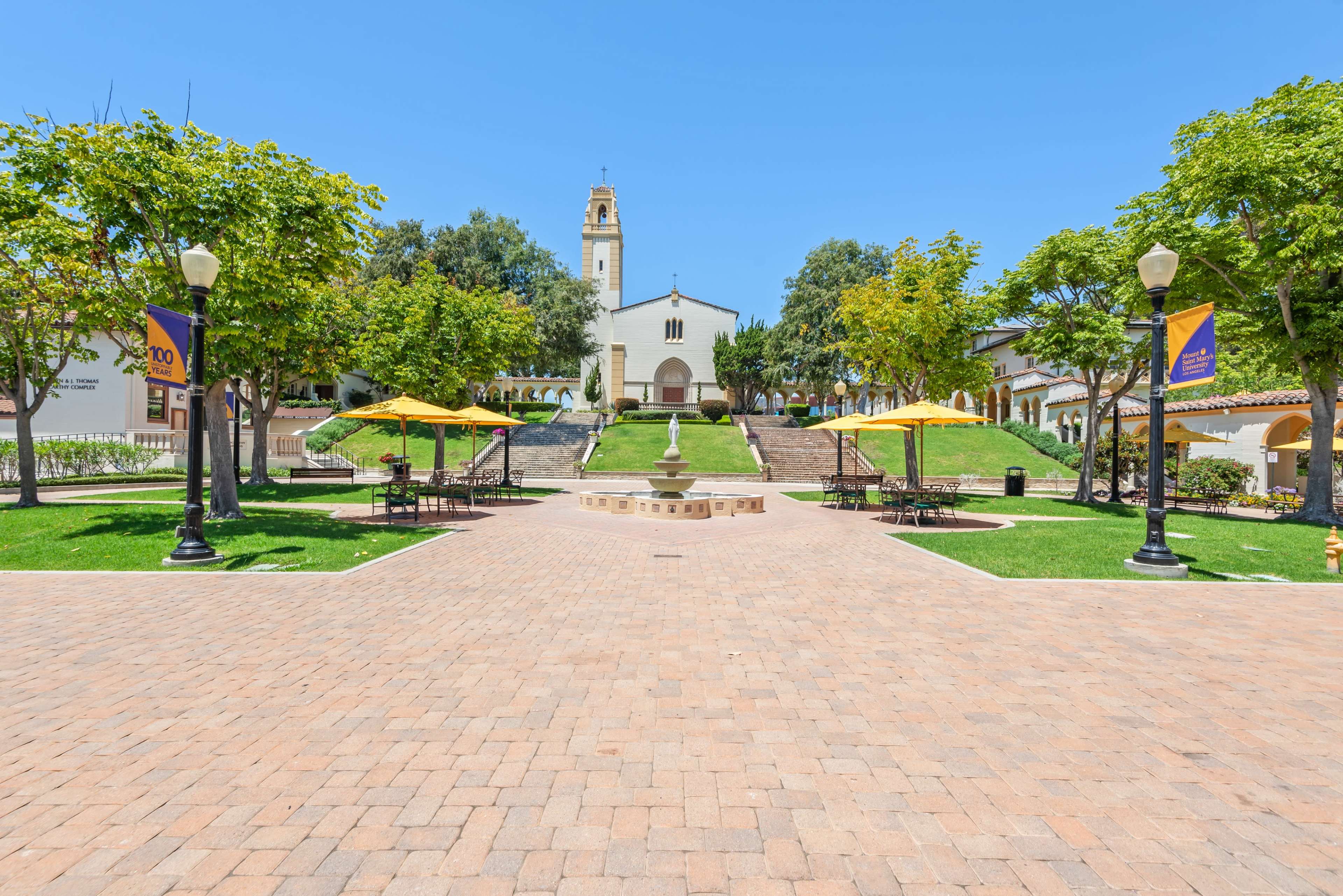 The image shows a sunny courtyard with yellow umbrellas over tables, leading towards a church with a tower in the background, surrounded by trees and green grass.