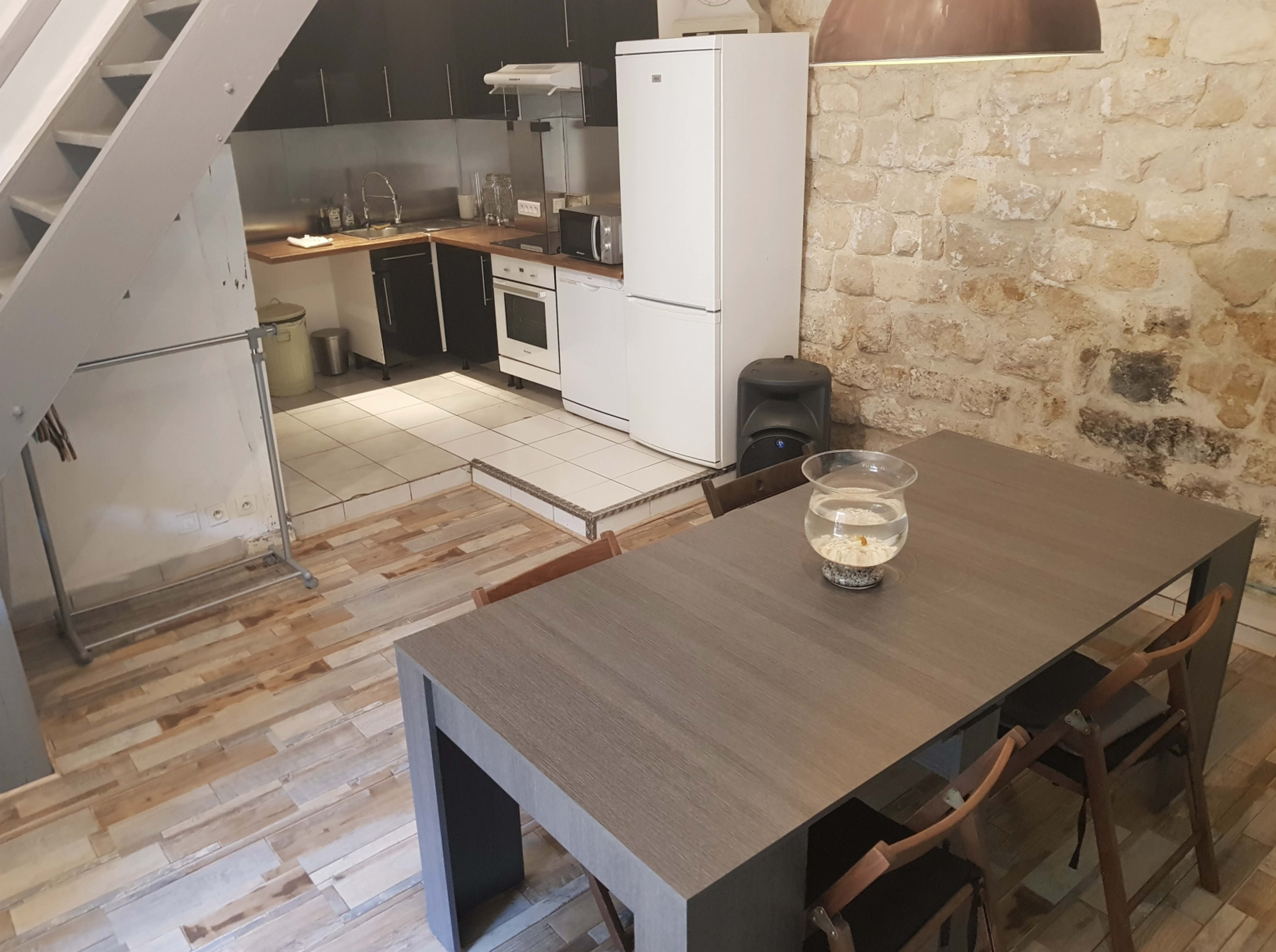 A modern dining area features a long table with chairs, adjacent to a small kitchen with appliances and exposed stone walls.