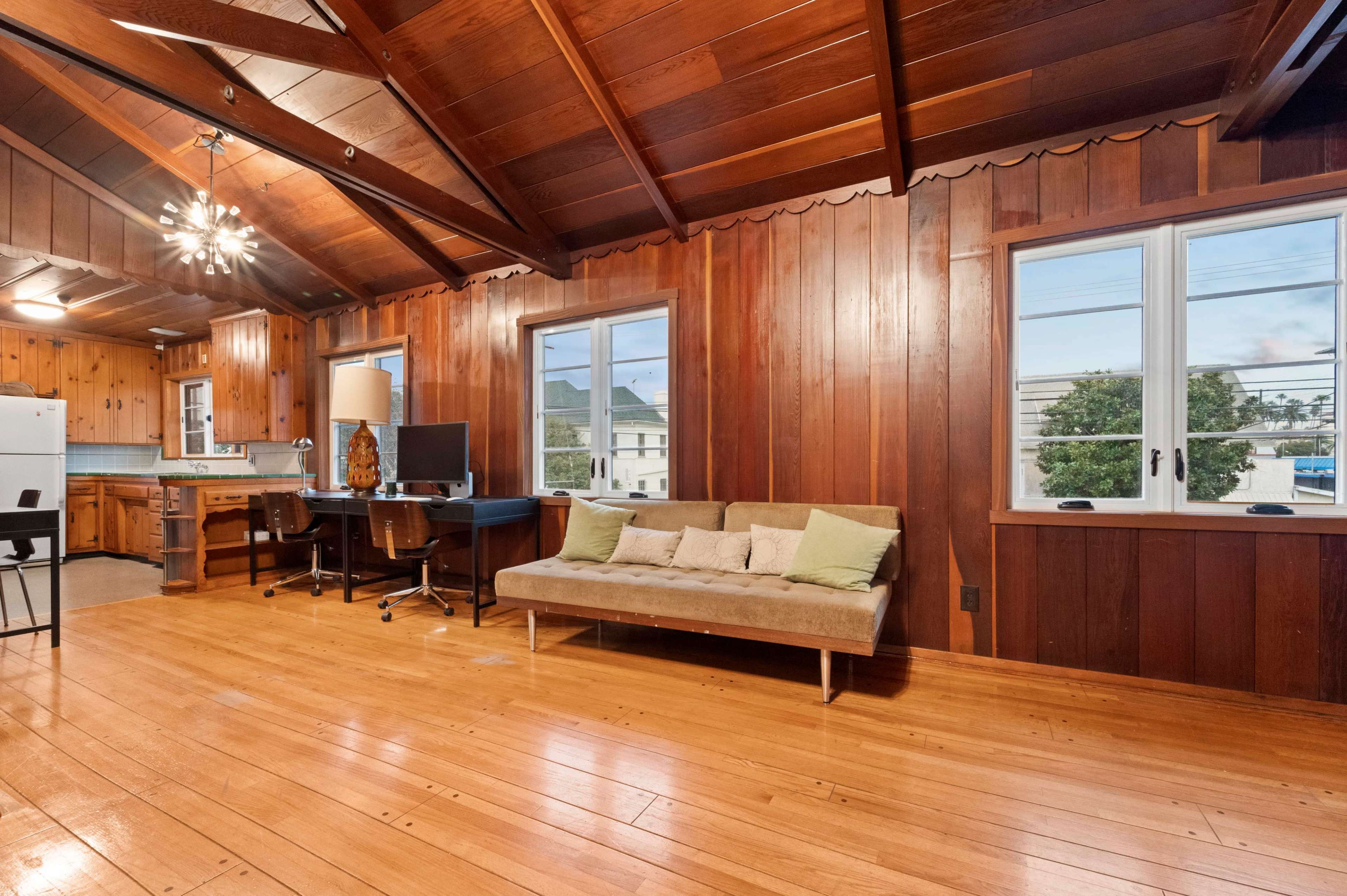 The room features a wooden interior with a sofa, desk, and computer, surrounded by large windows that allow natural light in.