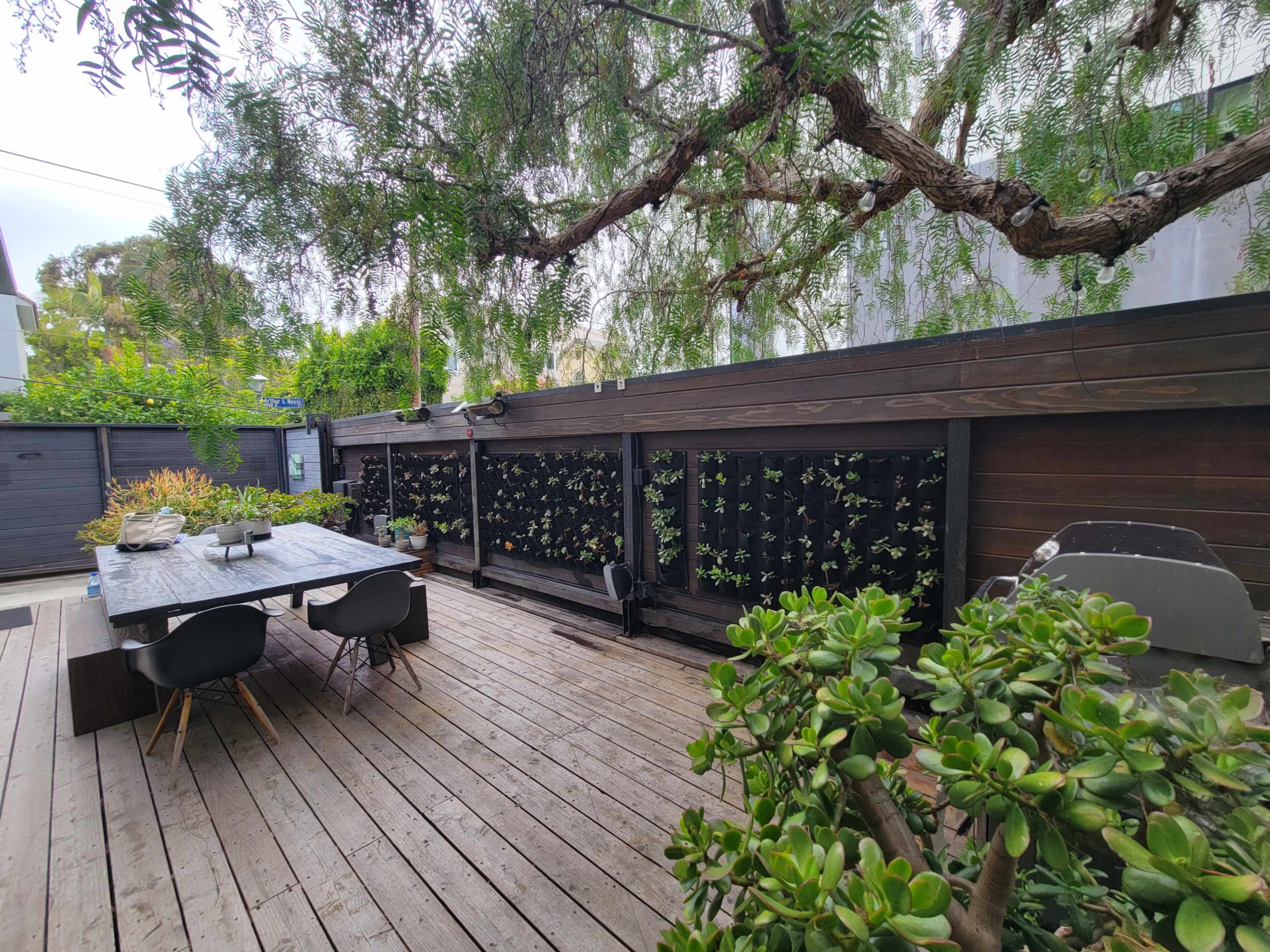 A wooden deck features a dining table and chairs, with a vertical garden on the wall and greenery surrounding the area.