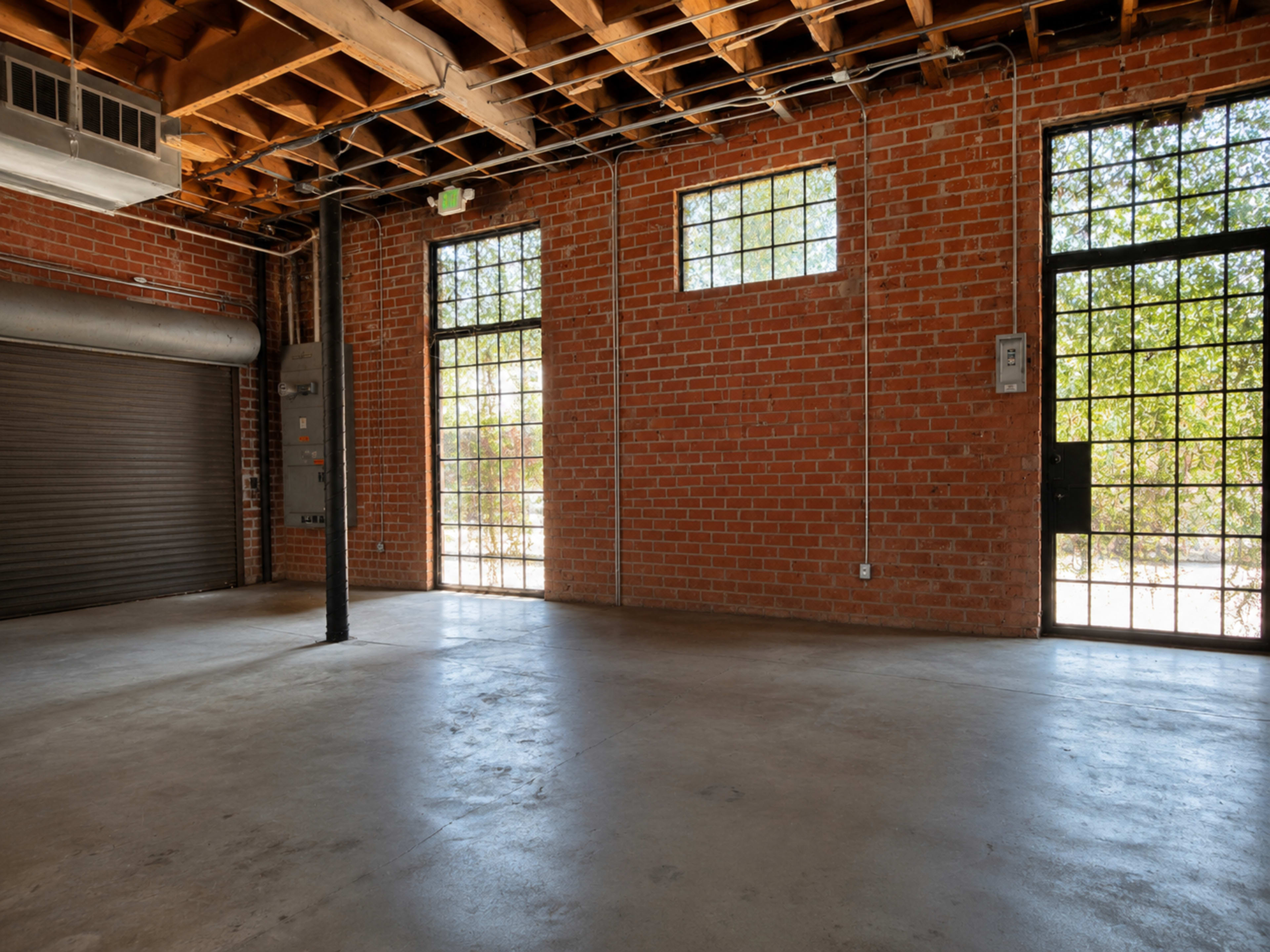 The image shows a spacious room with exposed brick walls, large windows on two sides, and a concrete floor.
