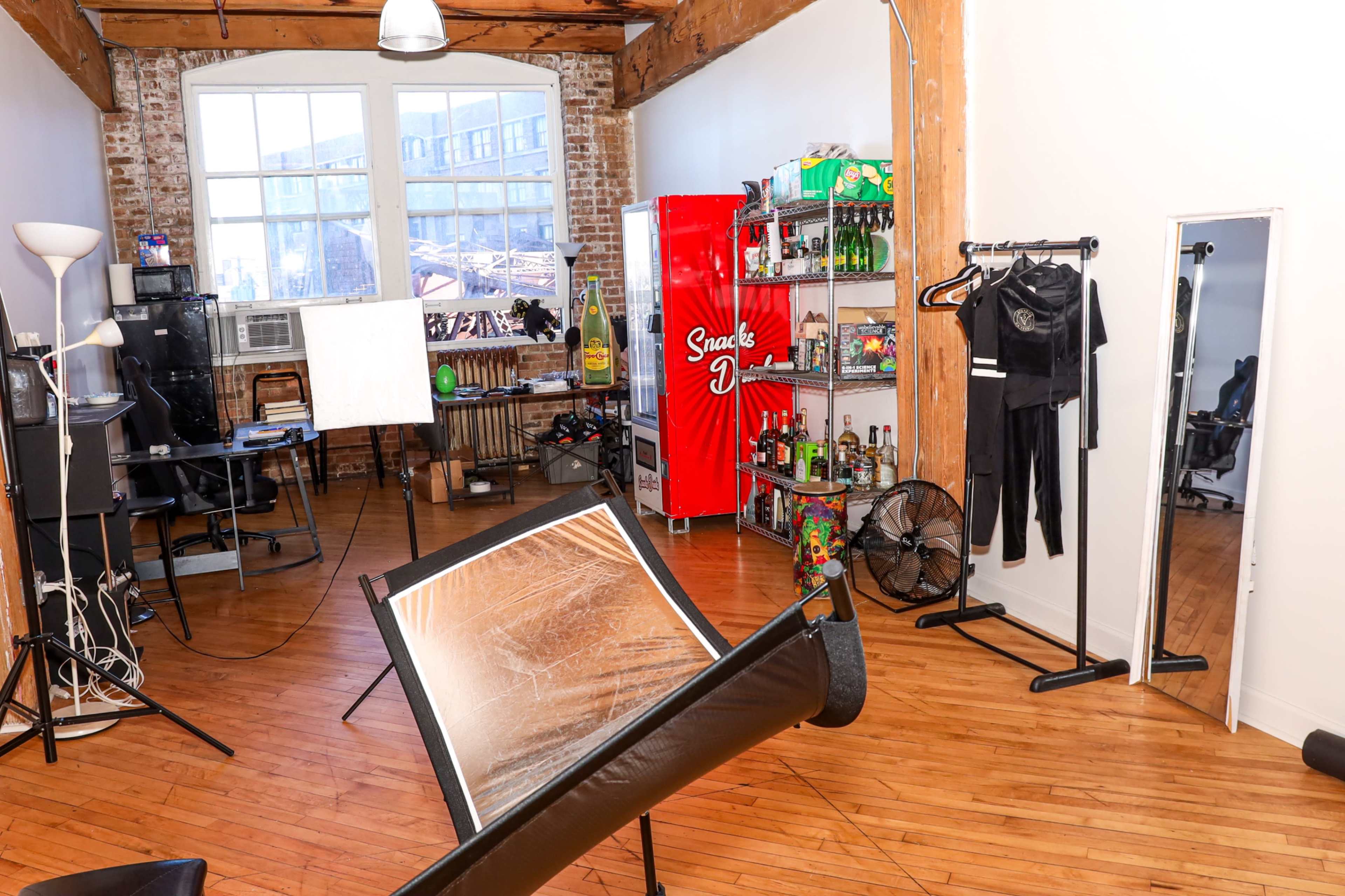 The image shows a spacious room with wooden floors, featuring a red beverage cooler, a clothing rack, and a desk with a computer setup.