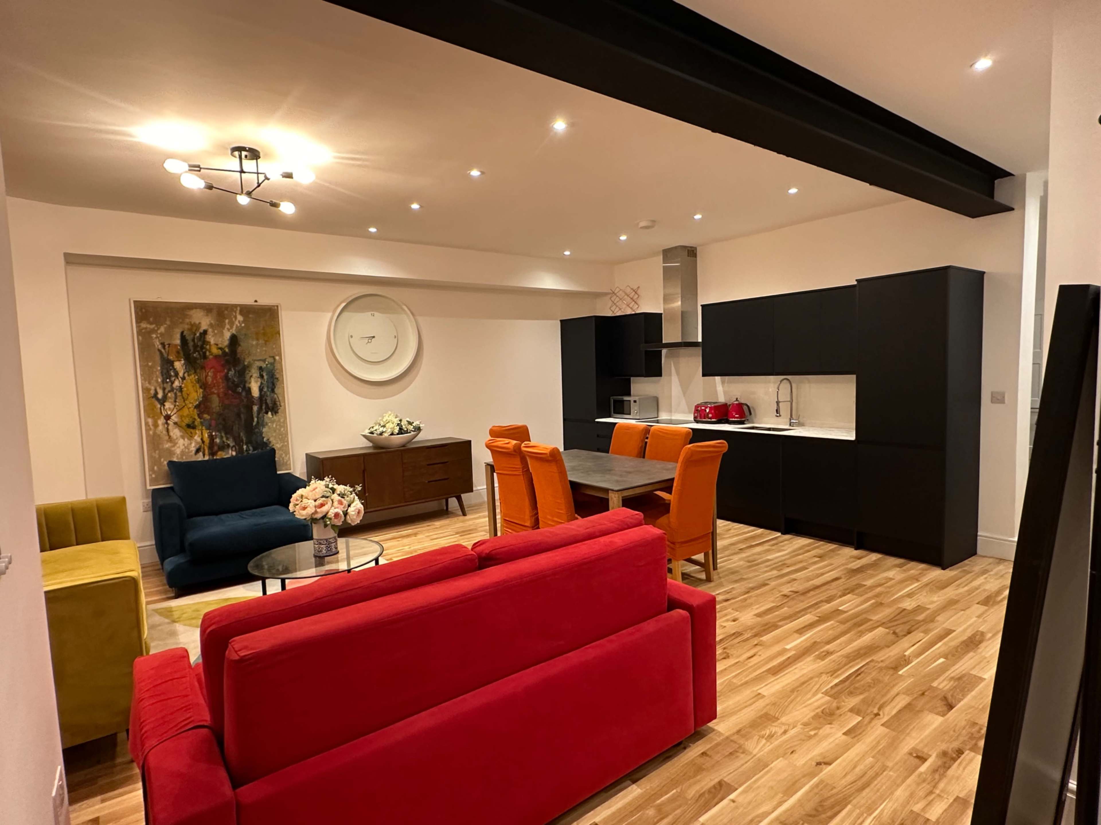 A modern living room and kitchen area with colorful furniture, including a red sofa and orange dining chairs, arranged around a coffee table.
