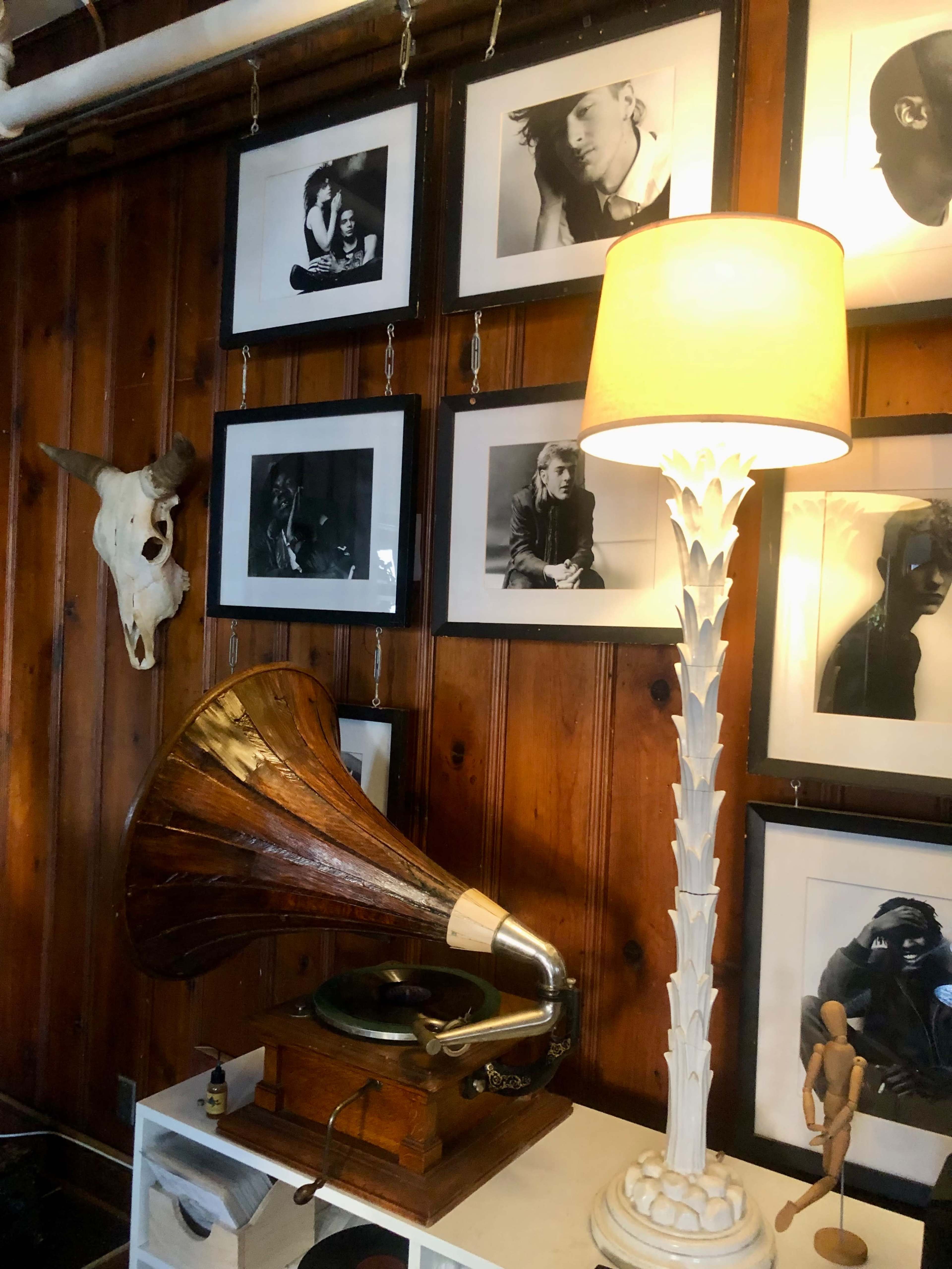 A vintage gramophone stands beside a tall lamp in a room with framed black-and-white photographs and a mounted skull on the wooden wall.