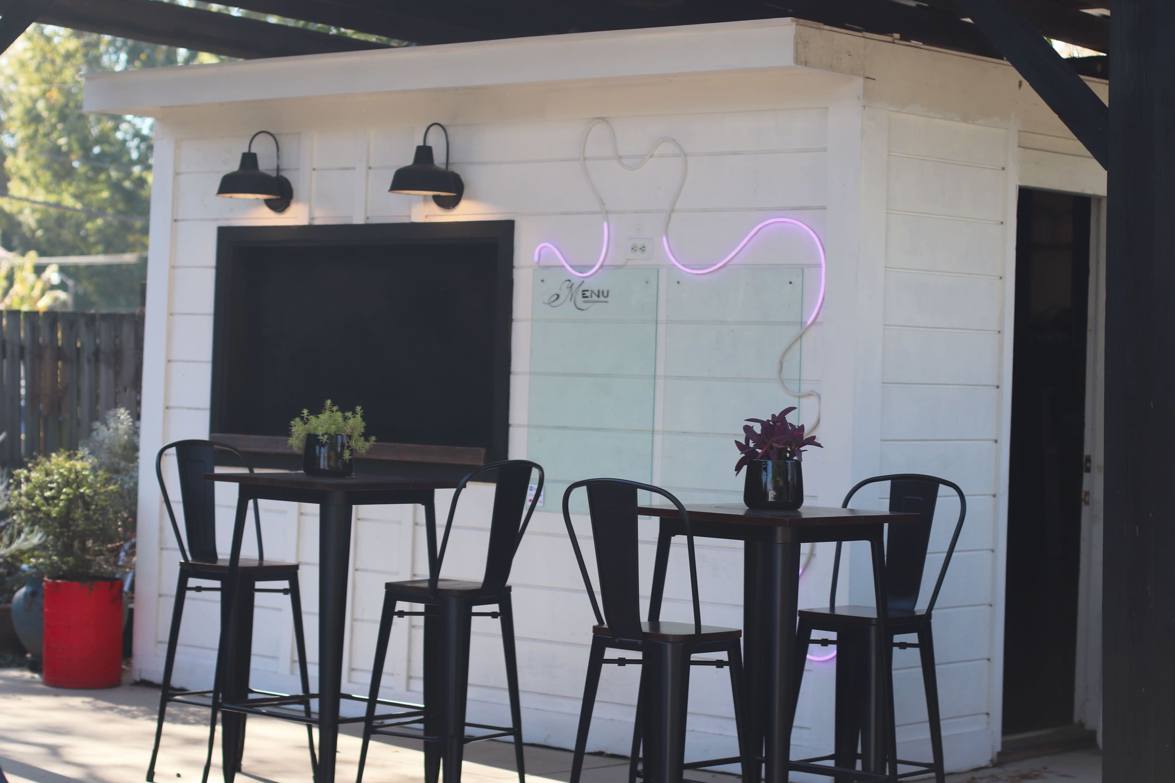 The image shows a modern outdoor bar setup with tall black chairs and a white wall featuring a chalkboard and neon lighting.