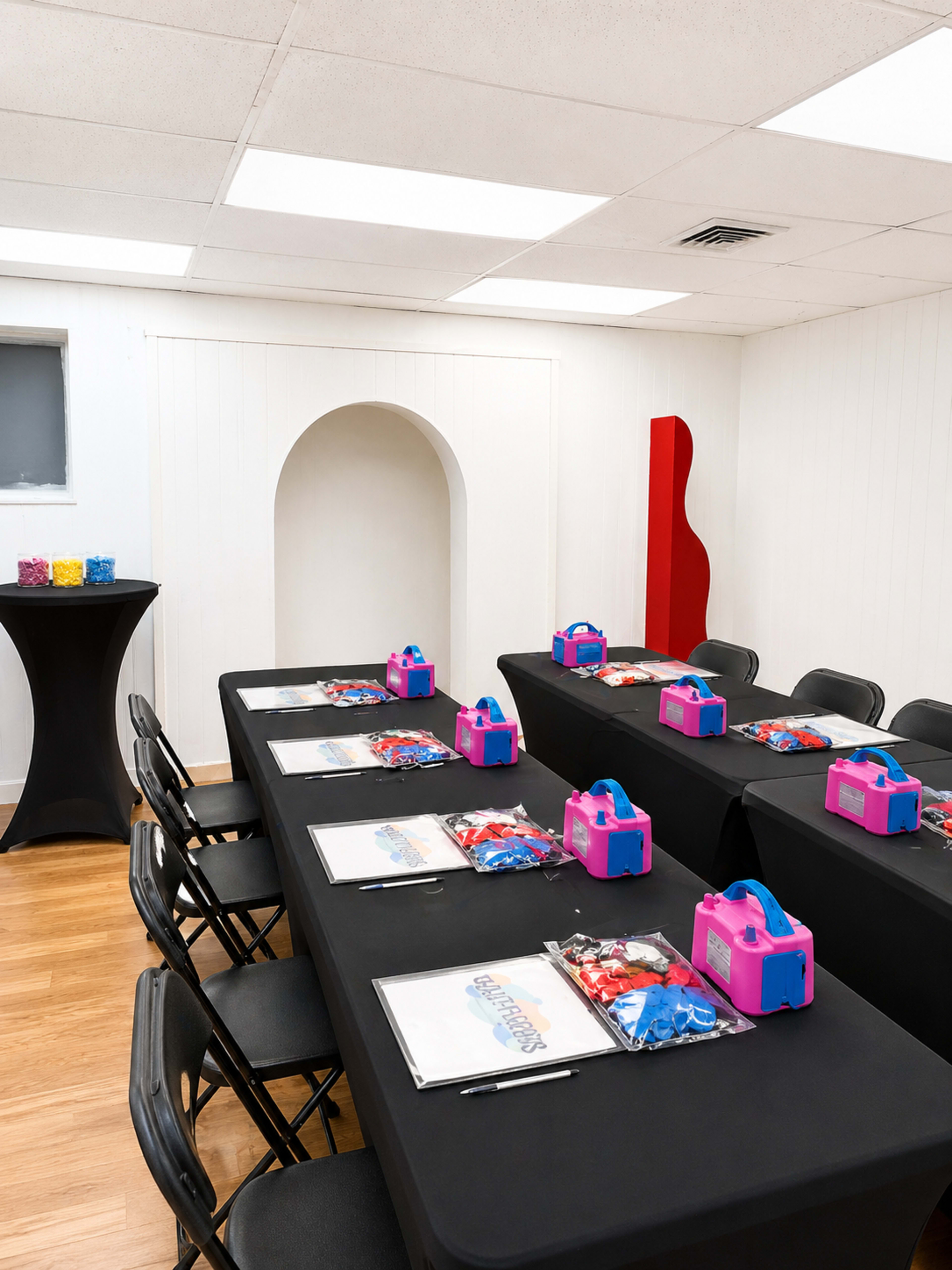 A conference room is set up with several black tables, each adorned with worksheets and colorful plastic containers, and there is a snack table in the corner.