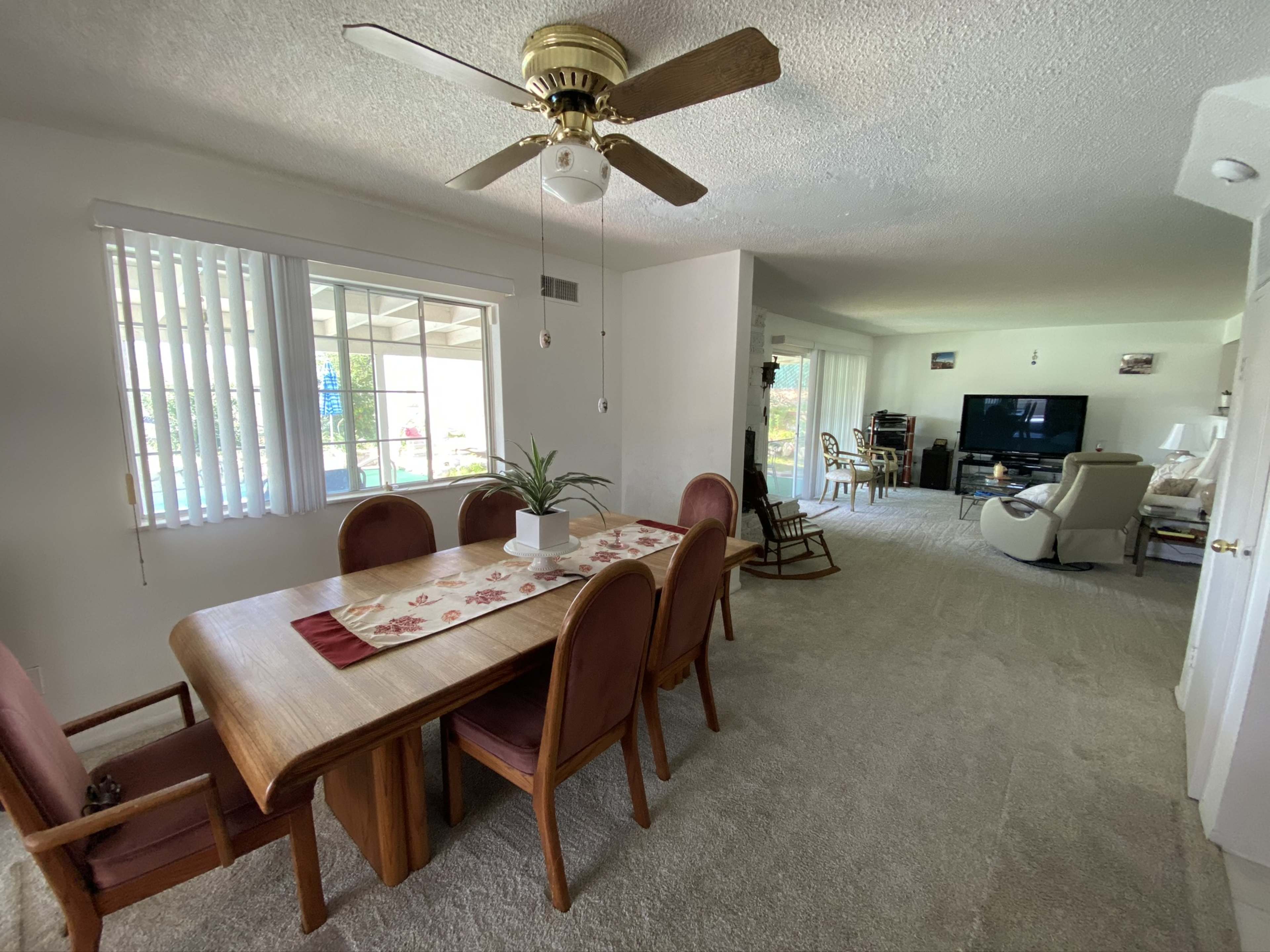 A well-lit living and dining area featuring a wooden dining table, chairs, and a television in the background.