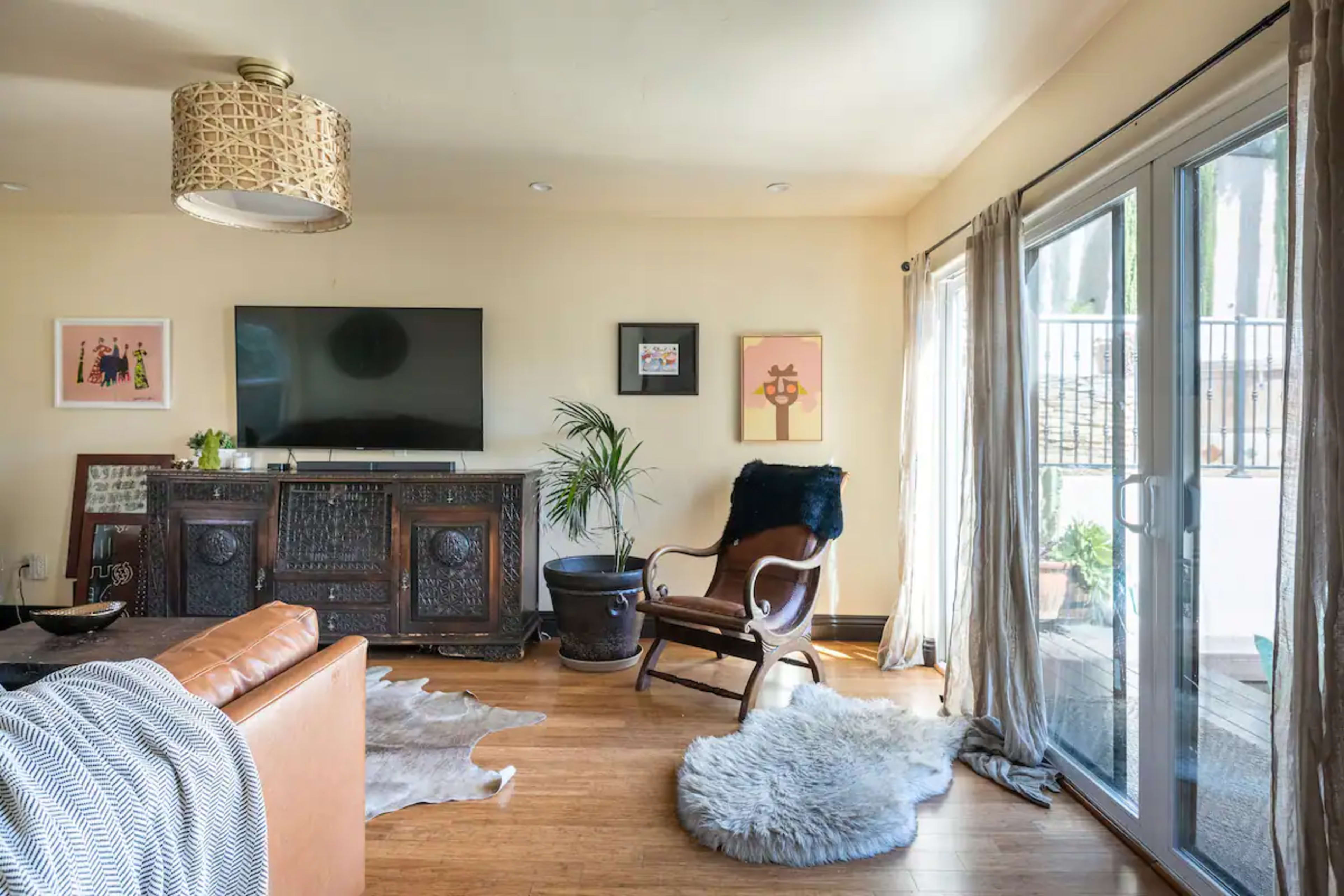 The living room features a wooden sideboard, a large television mounted on the wall, a leather chair, and sliding glass doors that open to an outdoor area.