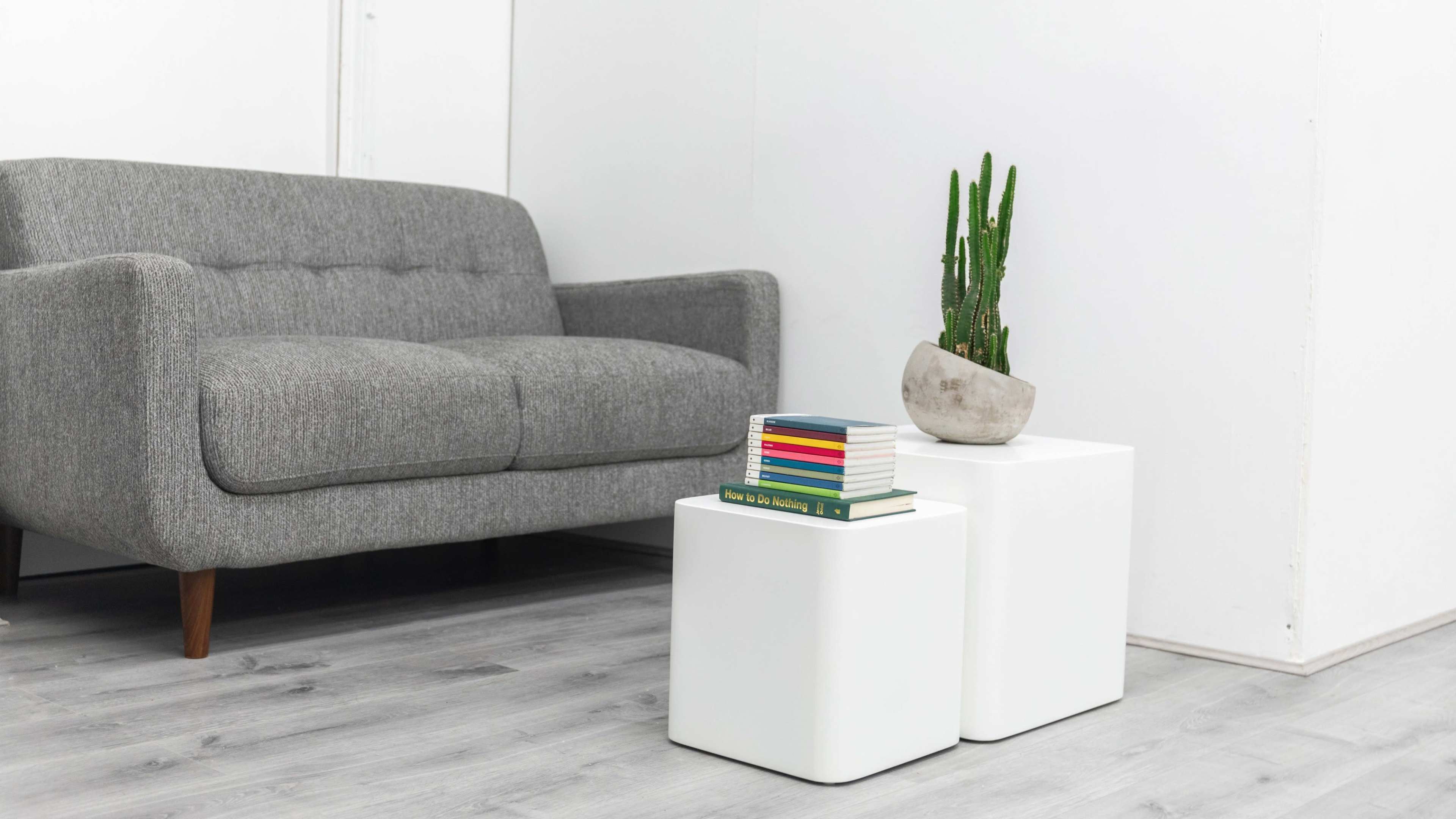 A gray couch sits beside two white tables stacked with colorful books and a potted cactus.