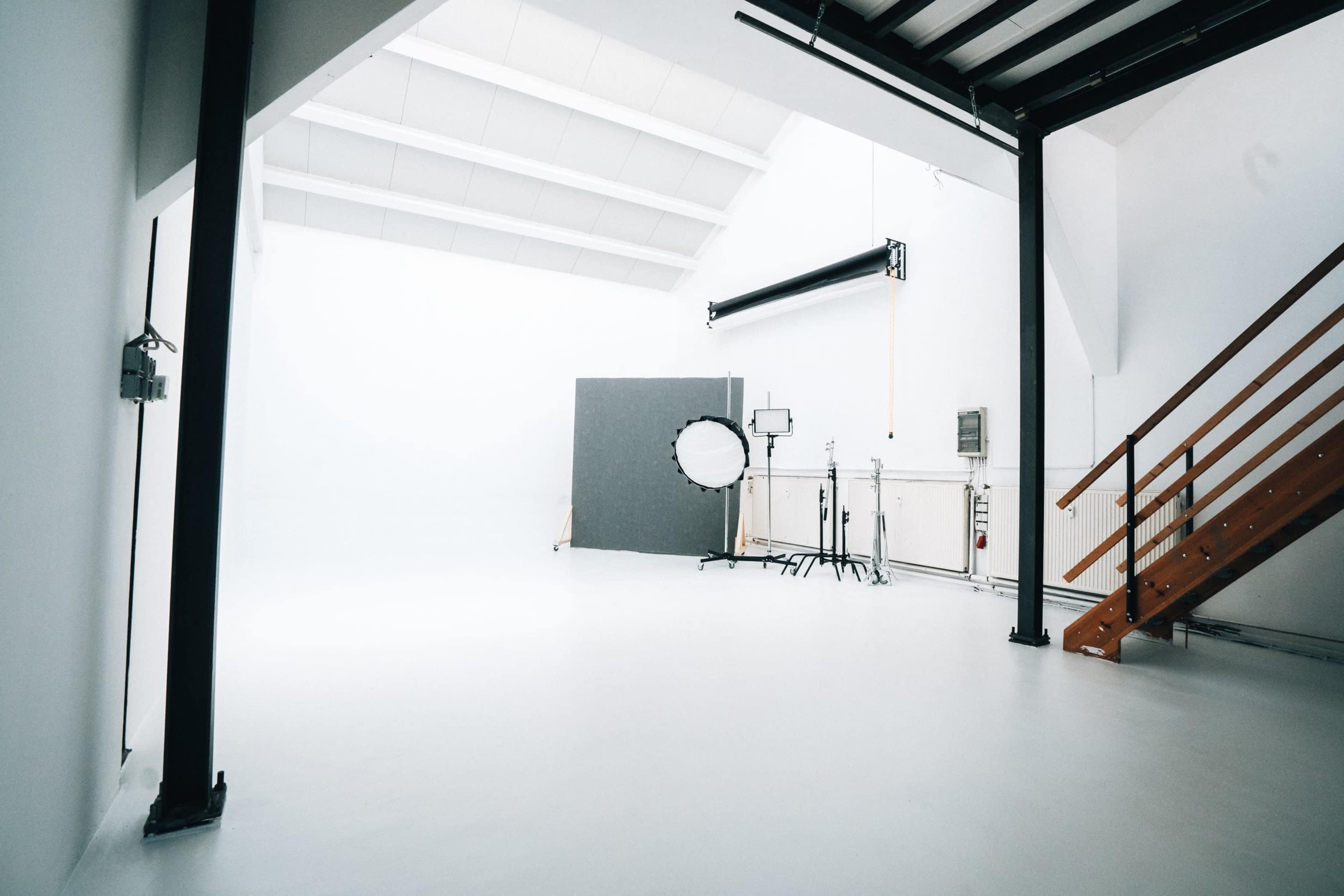A well-lit photography studio with a white background, a large round reflector on a stand, and a wooden staircase leading to a lofted area.