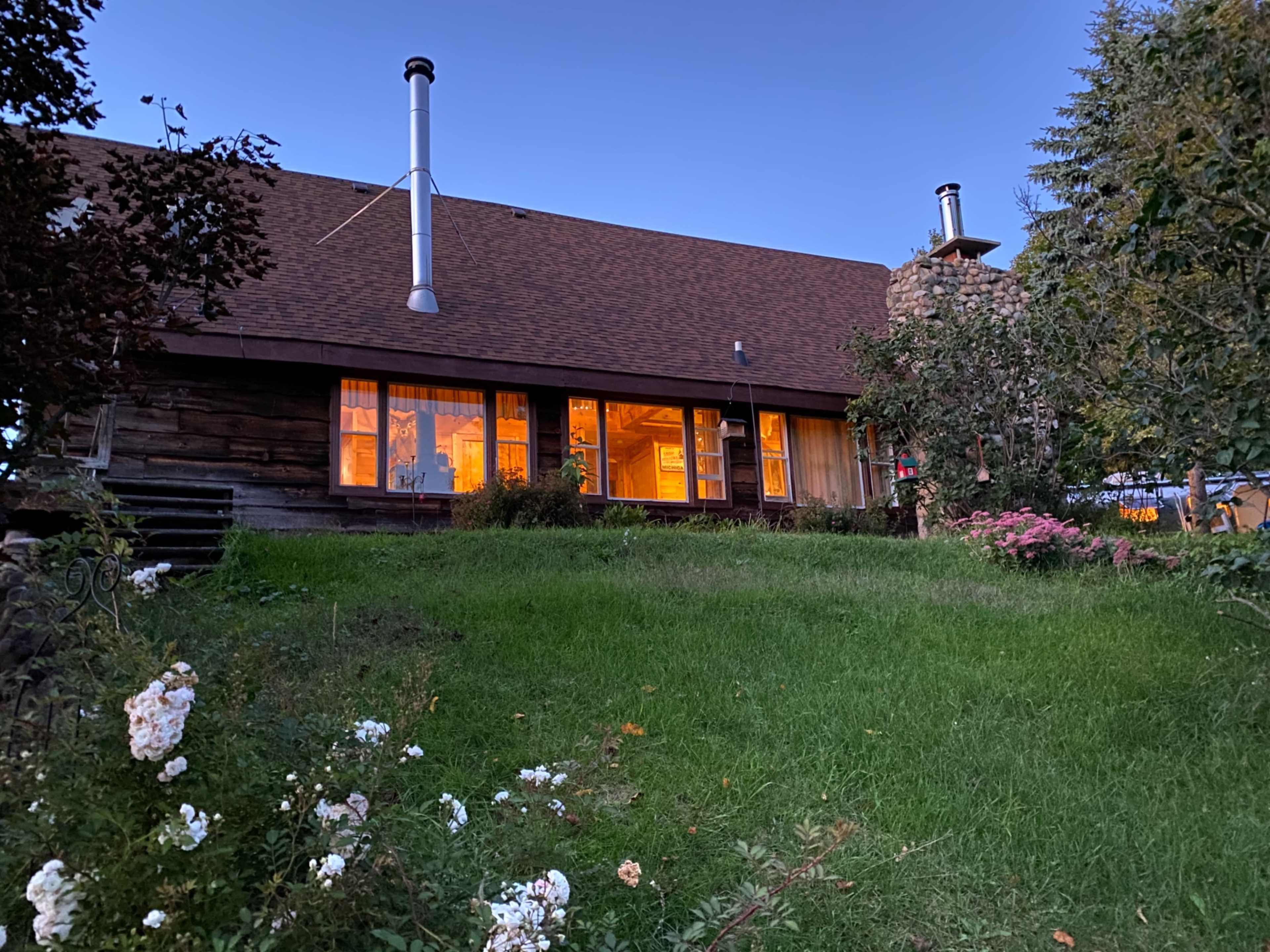 A wooden house with large windows and a stone chimney is set against a twilight sky, surrounded by a lawn and flowering plants.