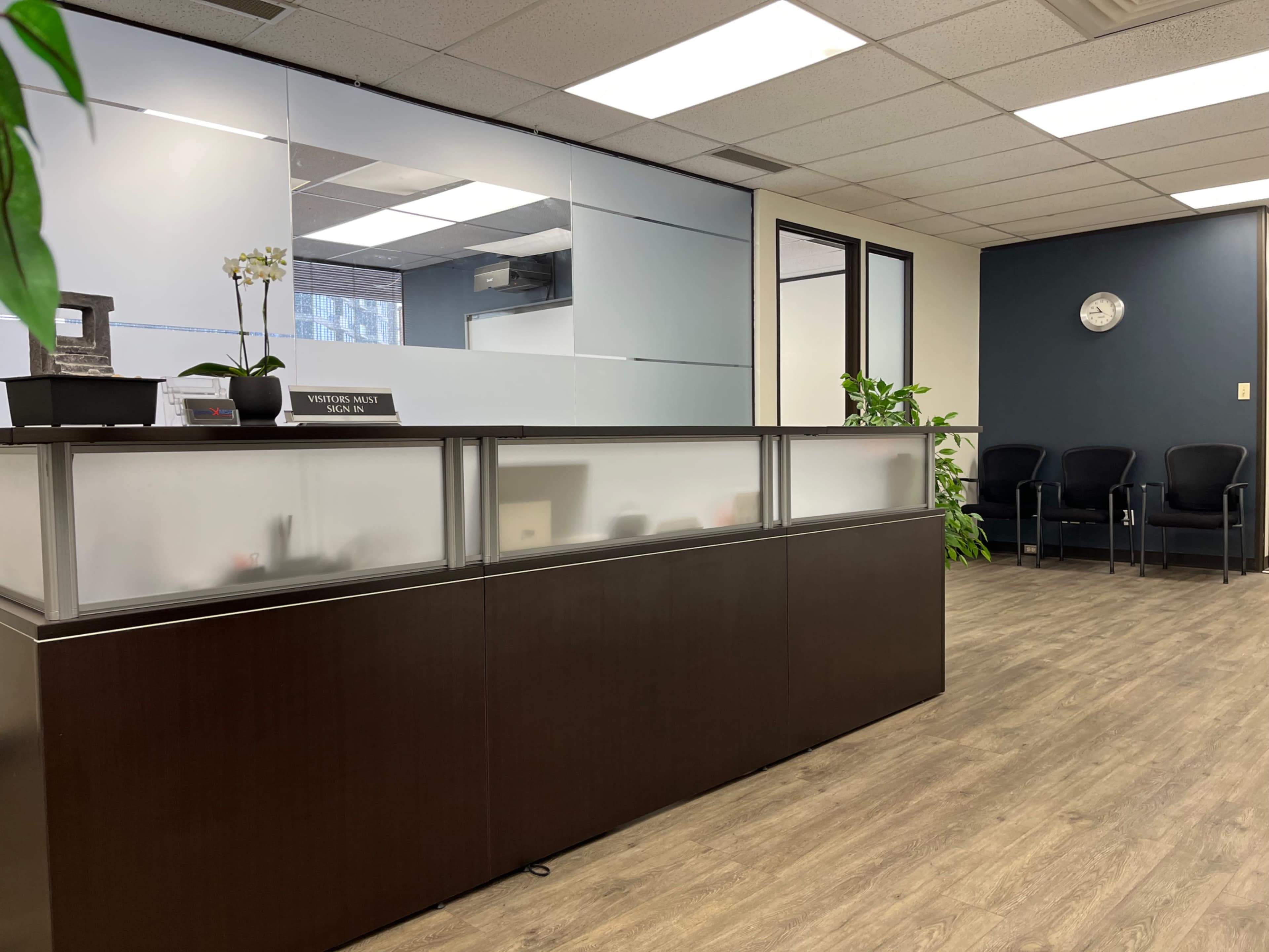 The image shows a modern office reception area with a low desk, seating for visitors, and large windows allowing natural light.