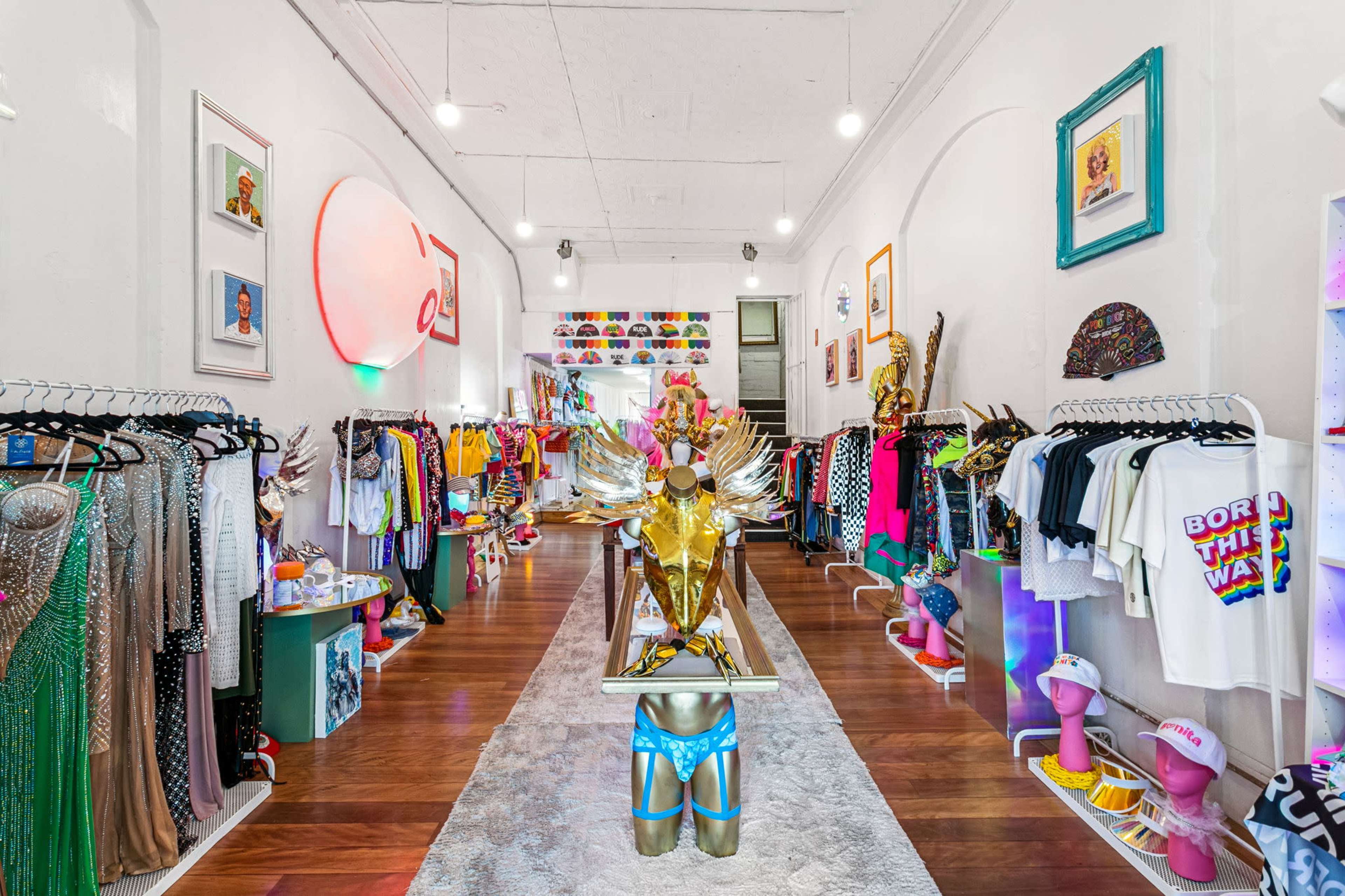 The image shows a colorful boutique filled with a variety of clothing and accessories displayed on racks, with a central decorative mannequin and mirrored wall at the back.